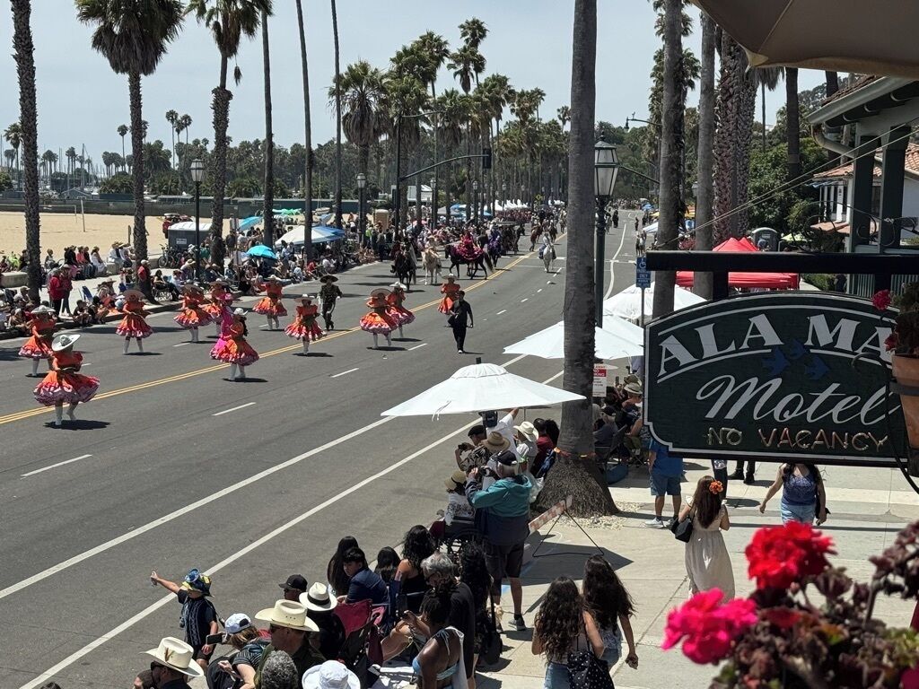 A street parade with performers in traditional costumes. A motel sign is visible in the foreground.