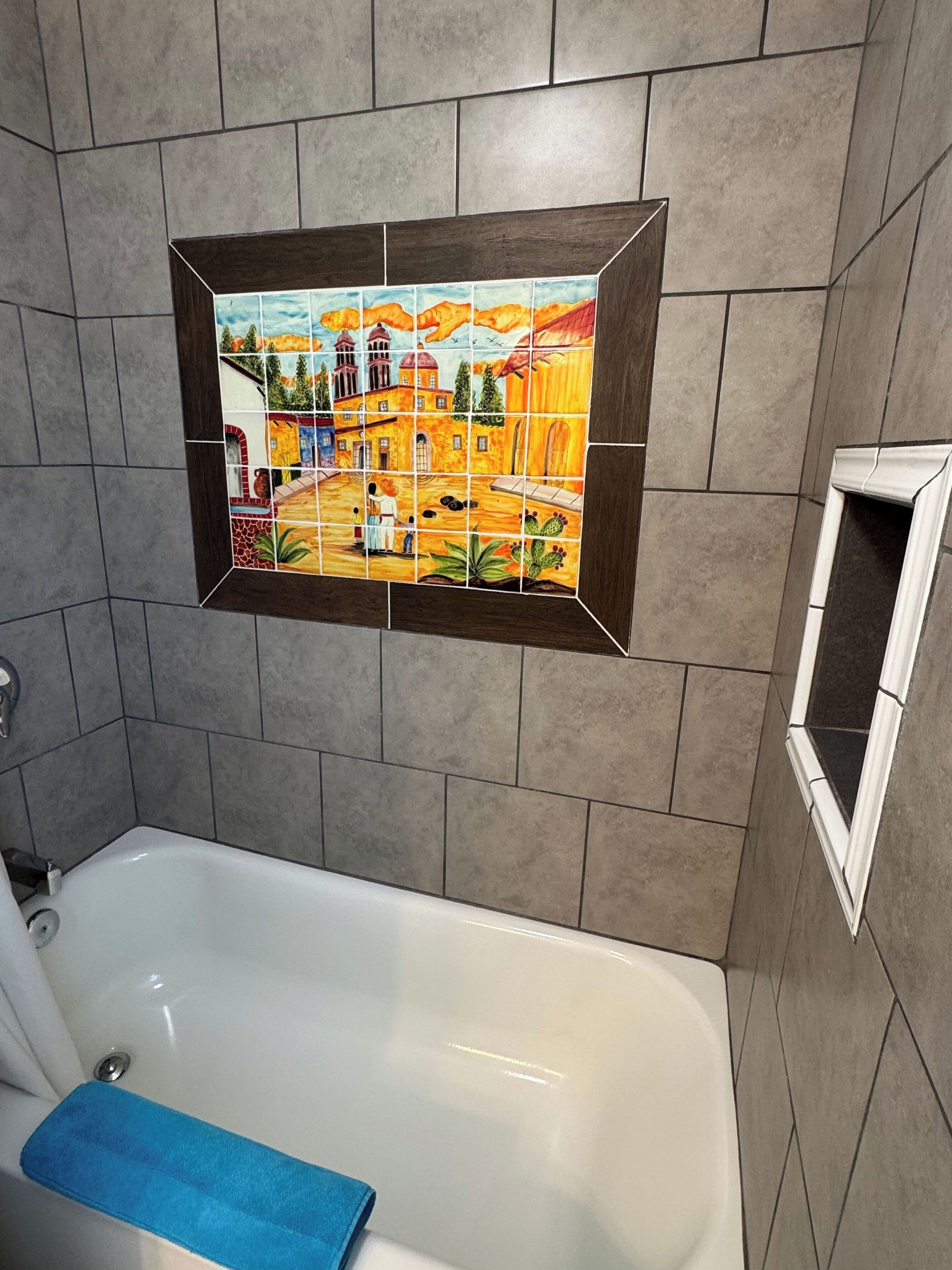 A bathroom with a white bathtub, a blue towel, and a mosaic tile wall depicting a landscape scene.