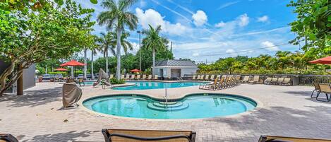A pool area with lounge chairs and umbrellas, surrounded by trees and a clear sky.