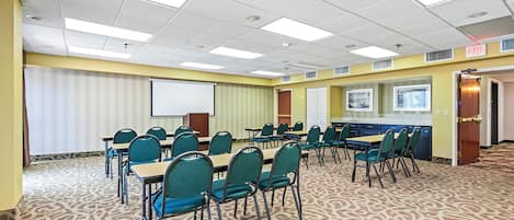 Un salón de conferencias con pantalla de proyector, mesas, sillas y piso alfombrado.