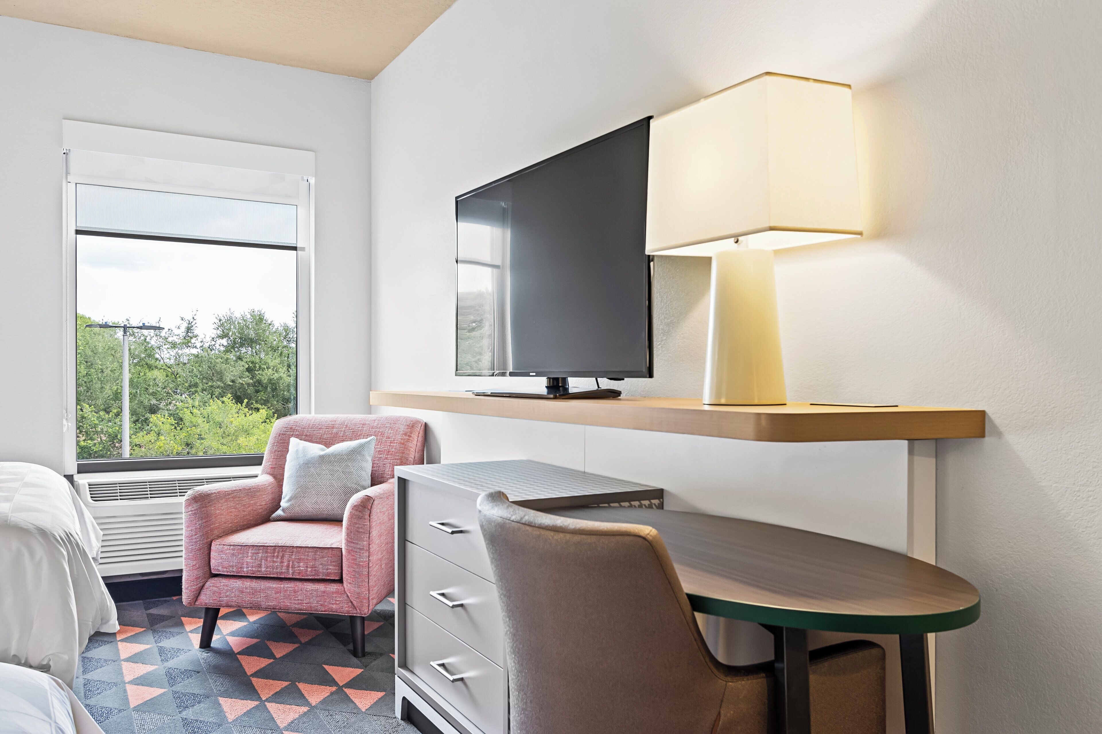 Een hotelkamer met een bed, bureau, stoel, televisie en een raam met uitzicht op bomen.