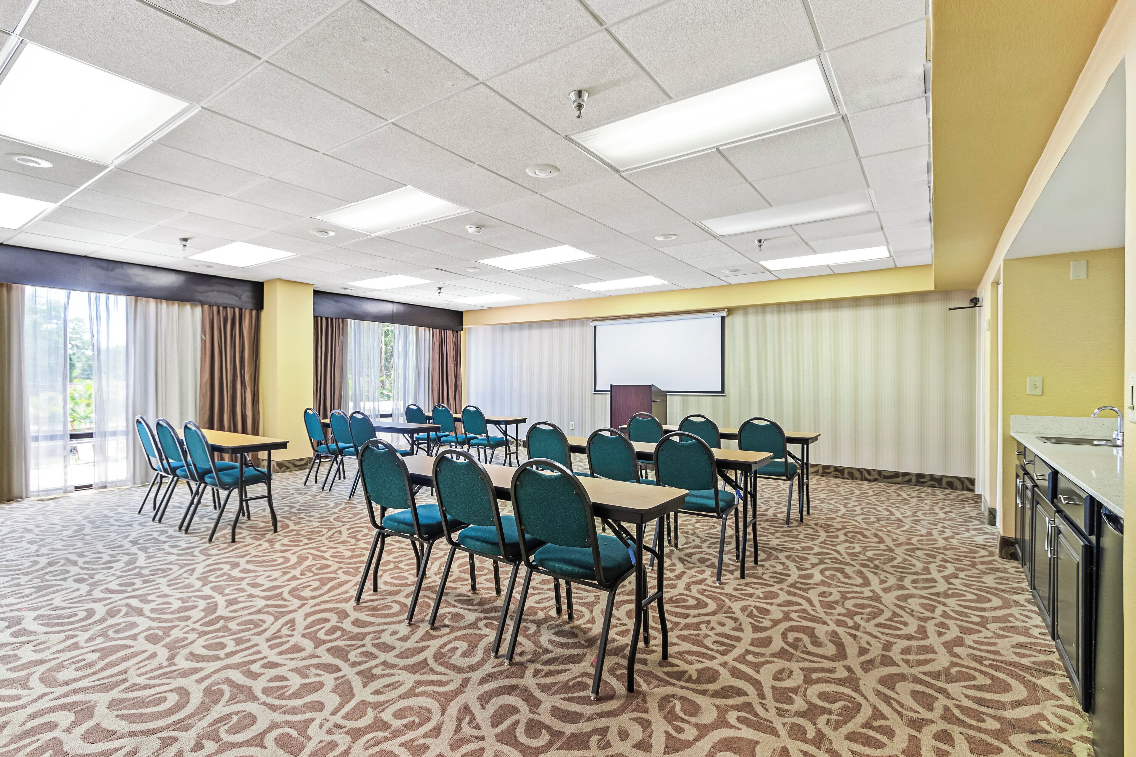 A conference room with a carpeted floor, tables, chairs, a projector screen, and a kitchenette.