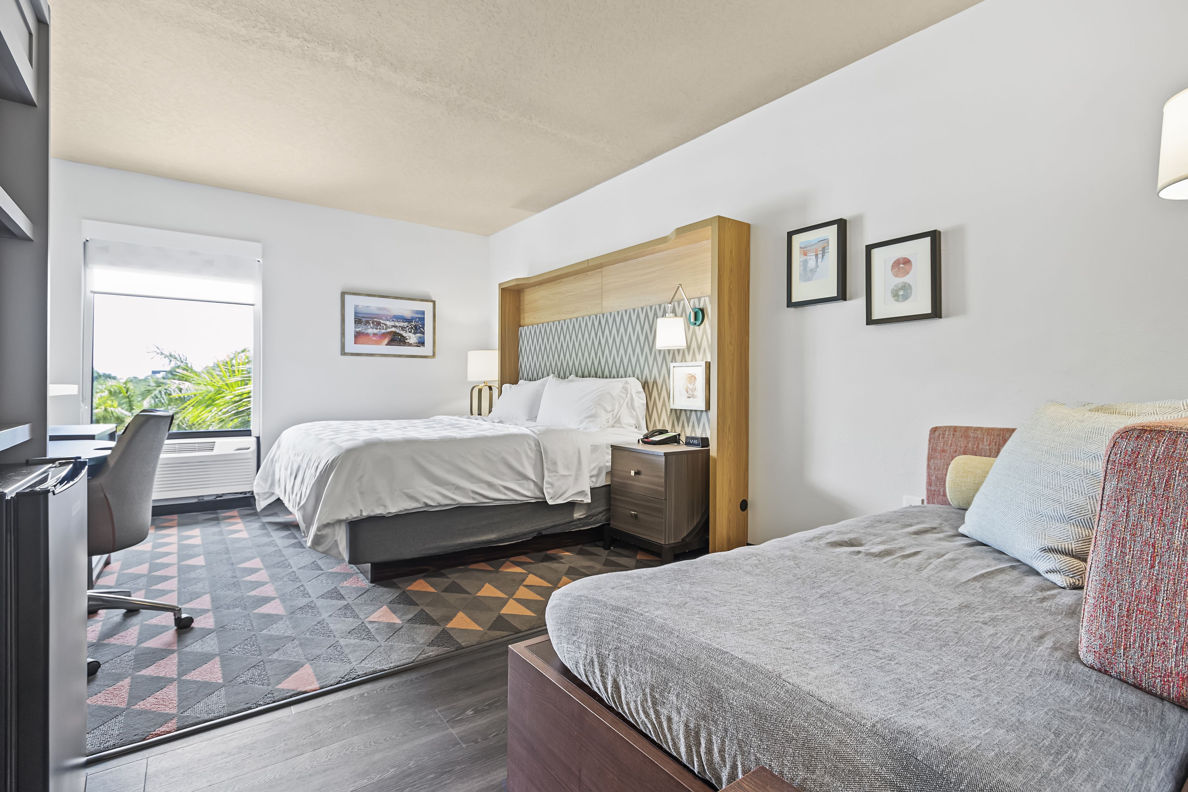 A hotel room with two beds, a desk, and a view of greenery outside.