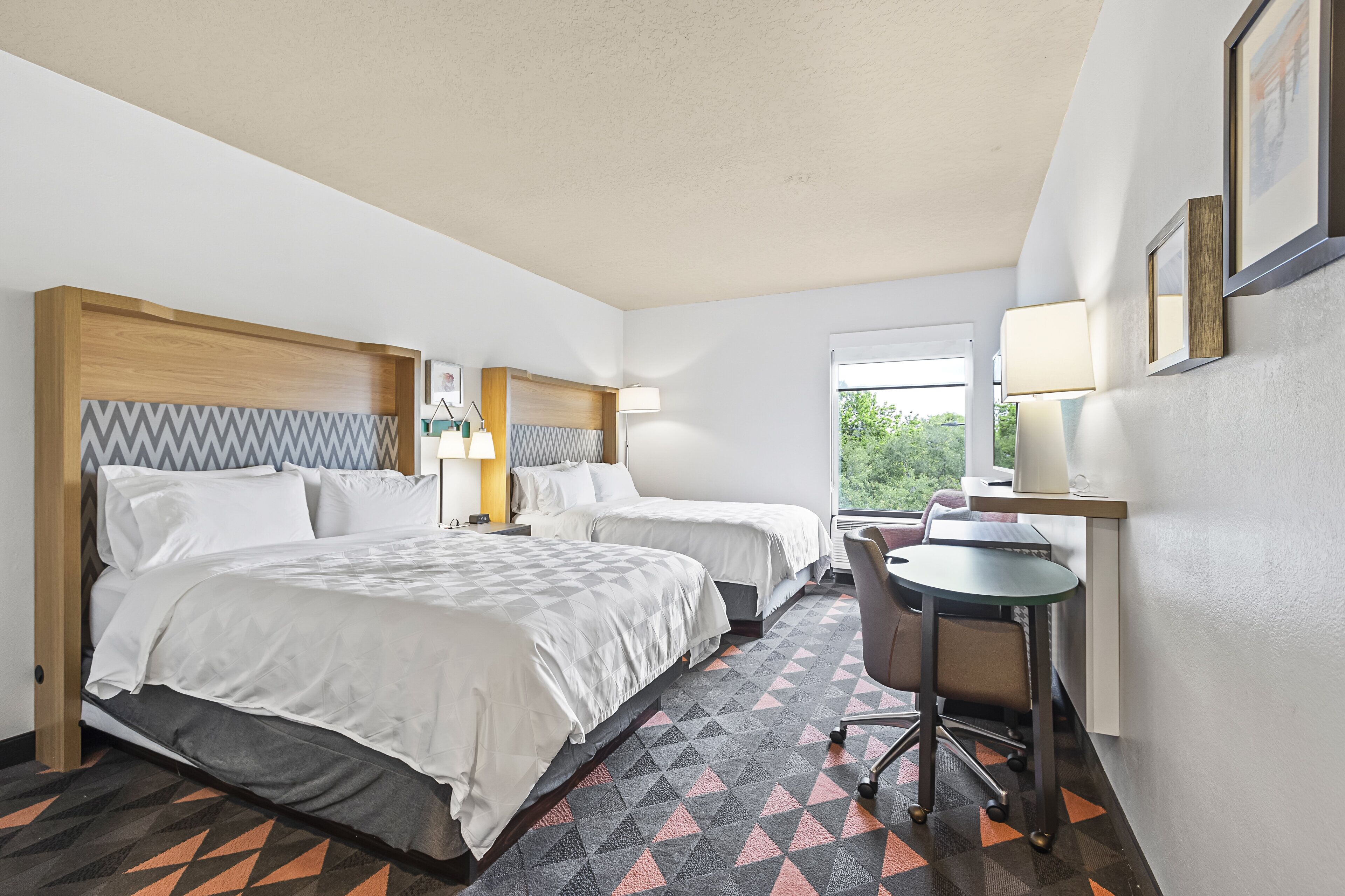 A hotel room with two beds, a desk, a chair, and a window with a view of greenery.