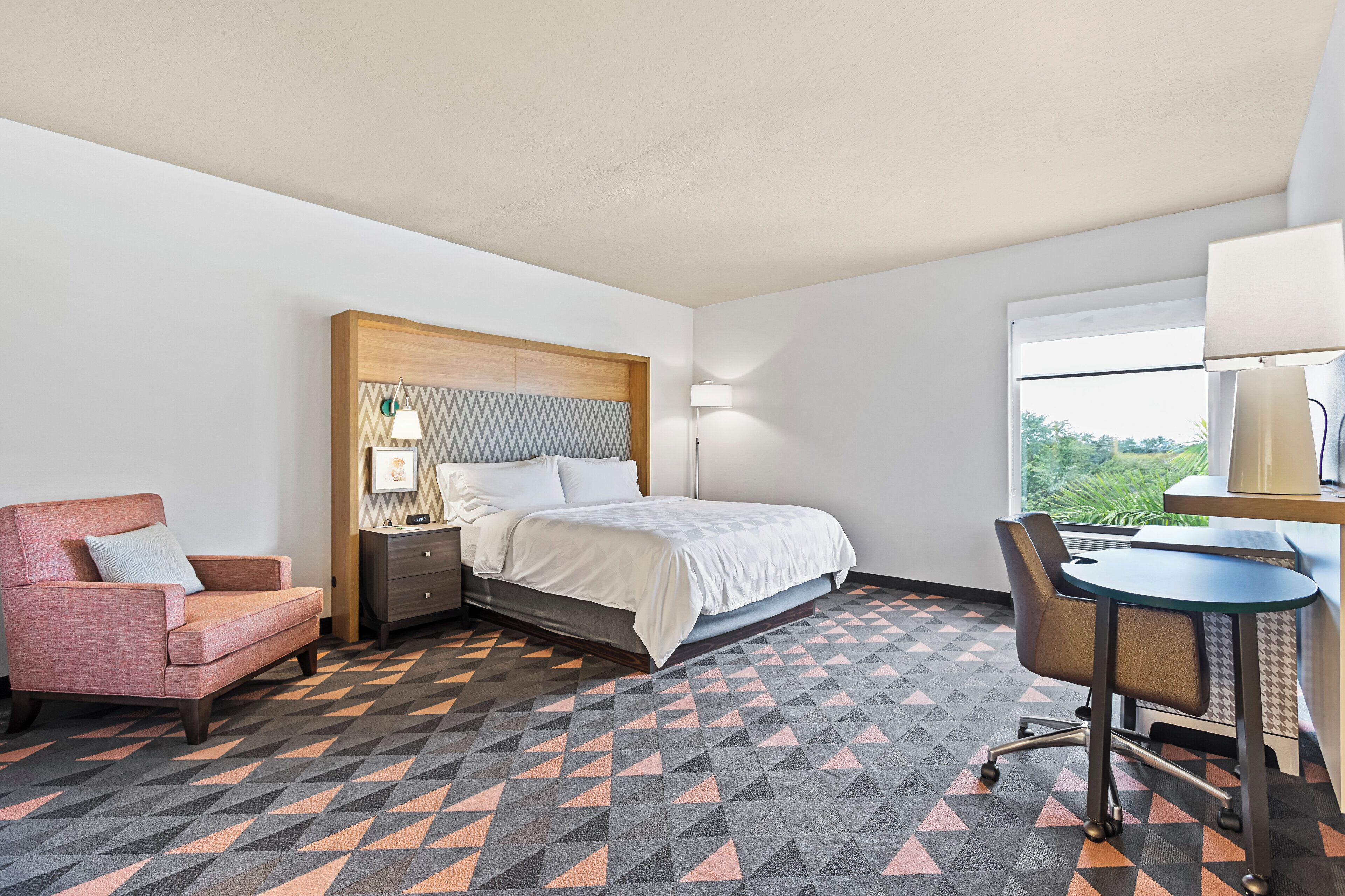 Una habitación de hotel con cama, escritorio, silla, mesita de noche y ventana.