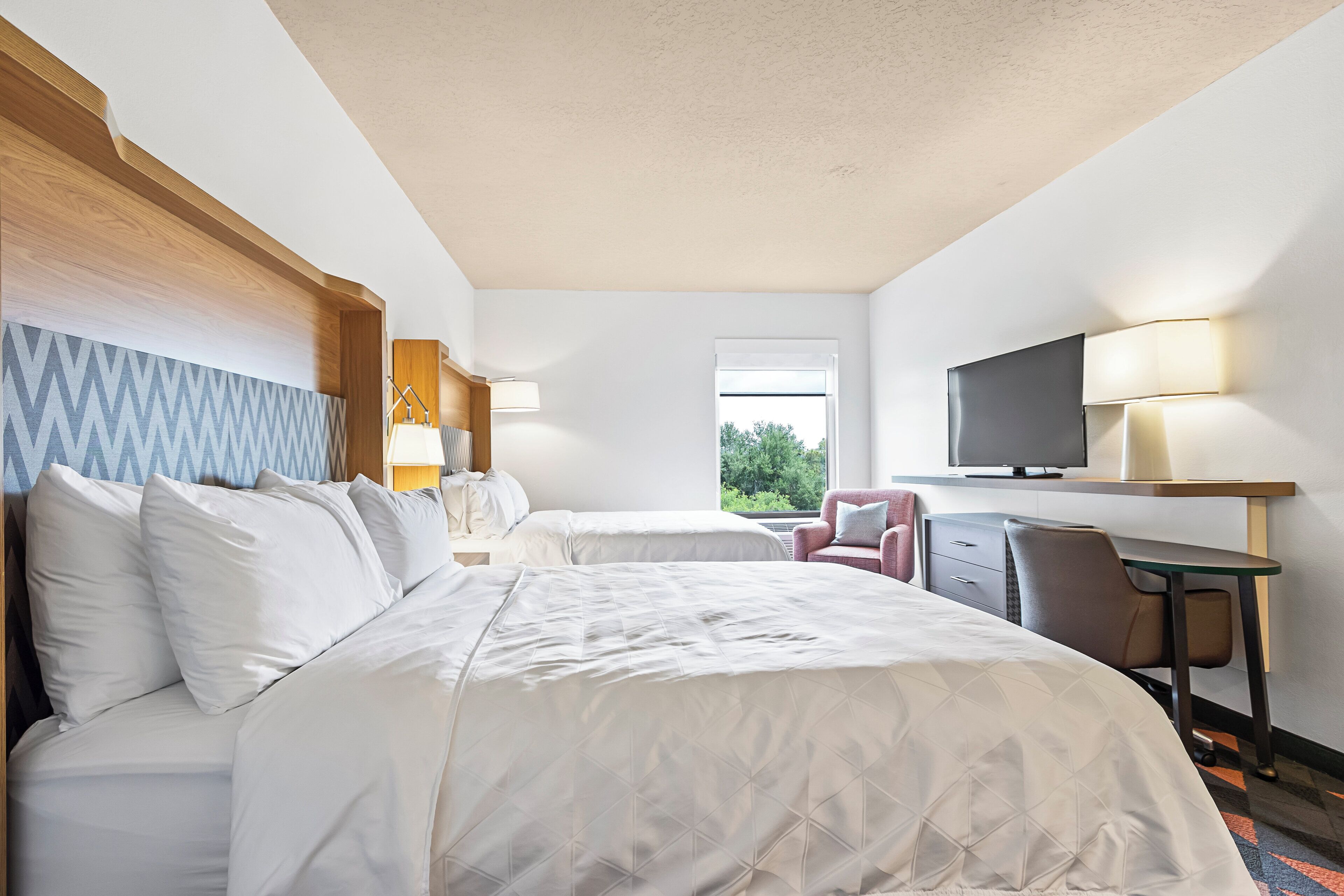 Ein Hotelzimmer mit zwei Betten, einem Schreibtisch, einem Stuhl, einem Fernseher und einem Fenster mit Blick auf Grünflächen.