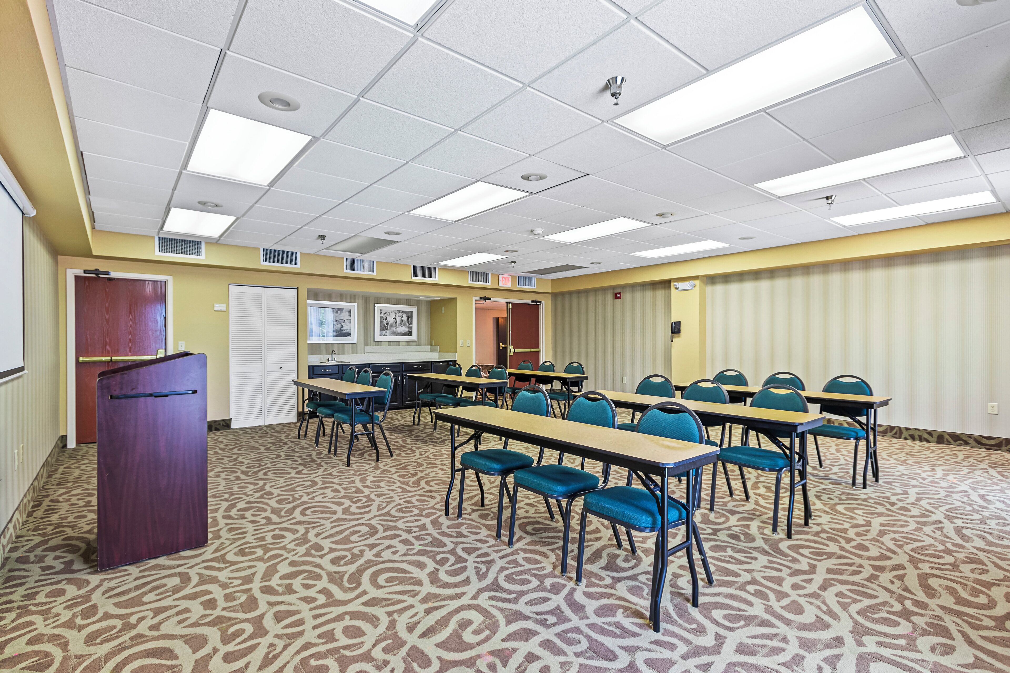 A conference room with a patterned carpet, rows of tables and chairs, a podium, and a kitchenette area.