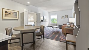A modern hotel room with a dining area, a living area with a sofa and armchair, a small round table, and a window with a view of greenery.