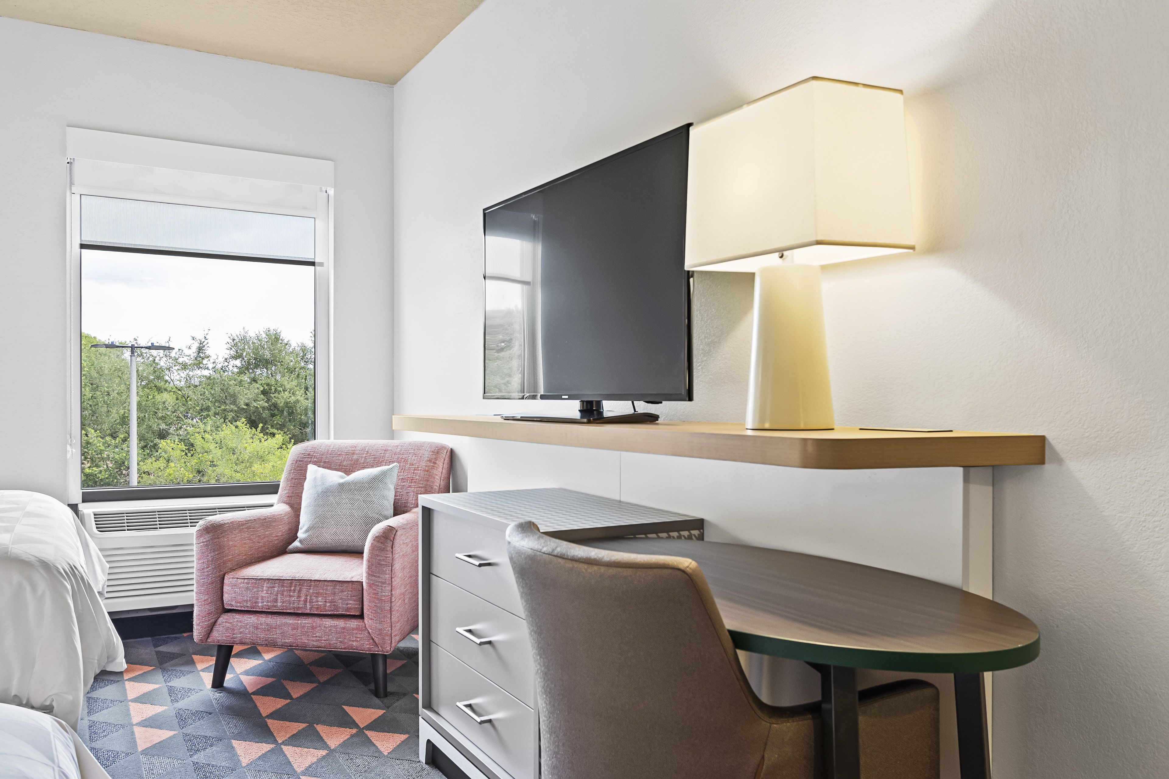 A hotel room with a bed, a desk with a lamp, a TV, and a chair.