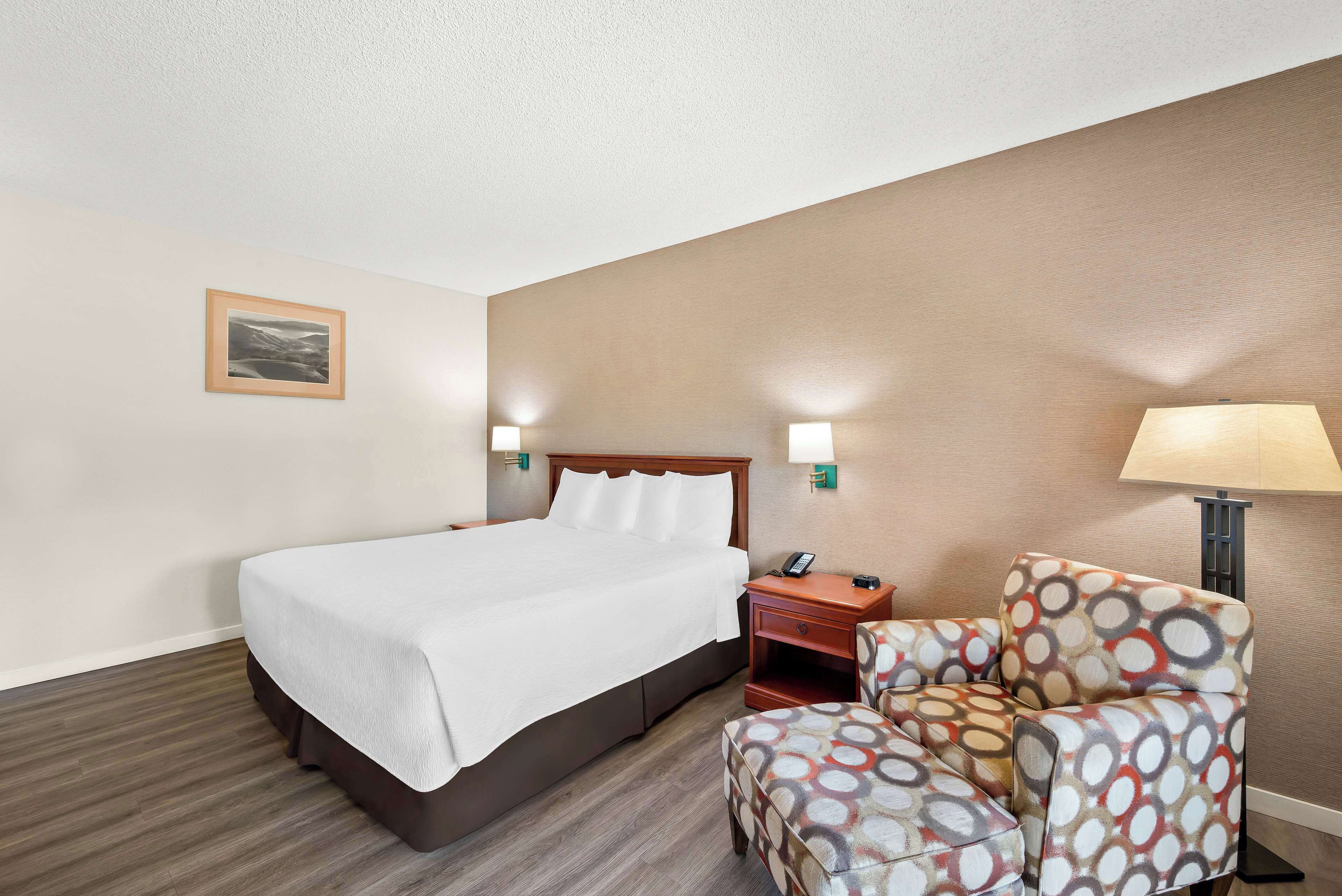 Quarto de hotel com cama, poltrona, mesa de cabeceira e abajur.