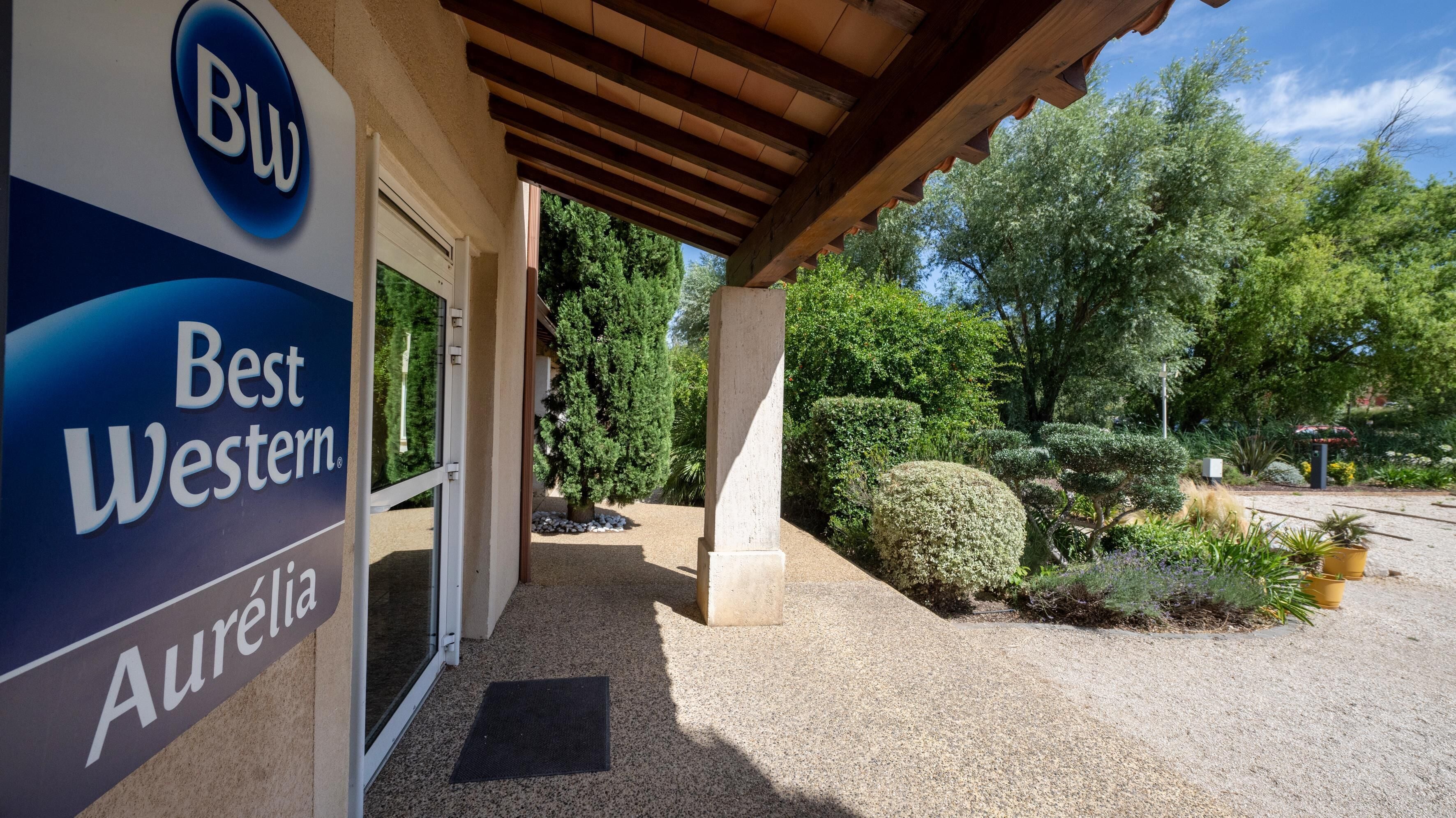 A Best Western hotel entrance with a covered patio area, a gravel pathway, and well-maintained landscaping.