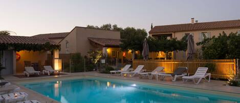 A swimming pool with lounge chairs and umbrellas, surrounded by buildings with tiled roofs and a wooden fence.