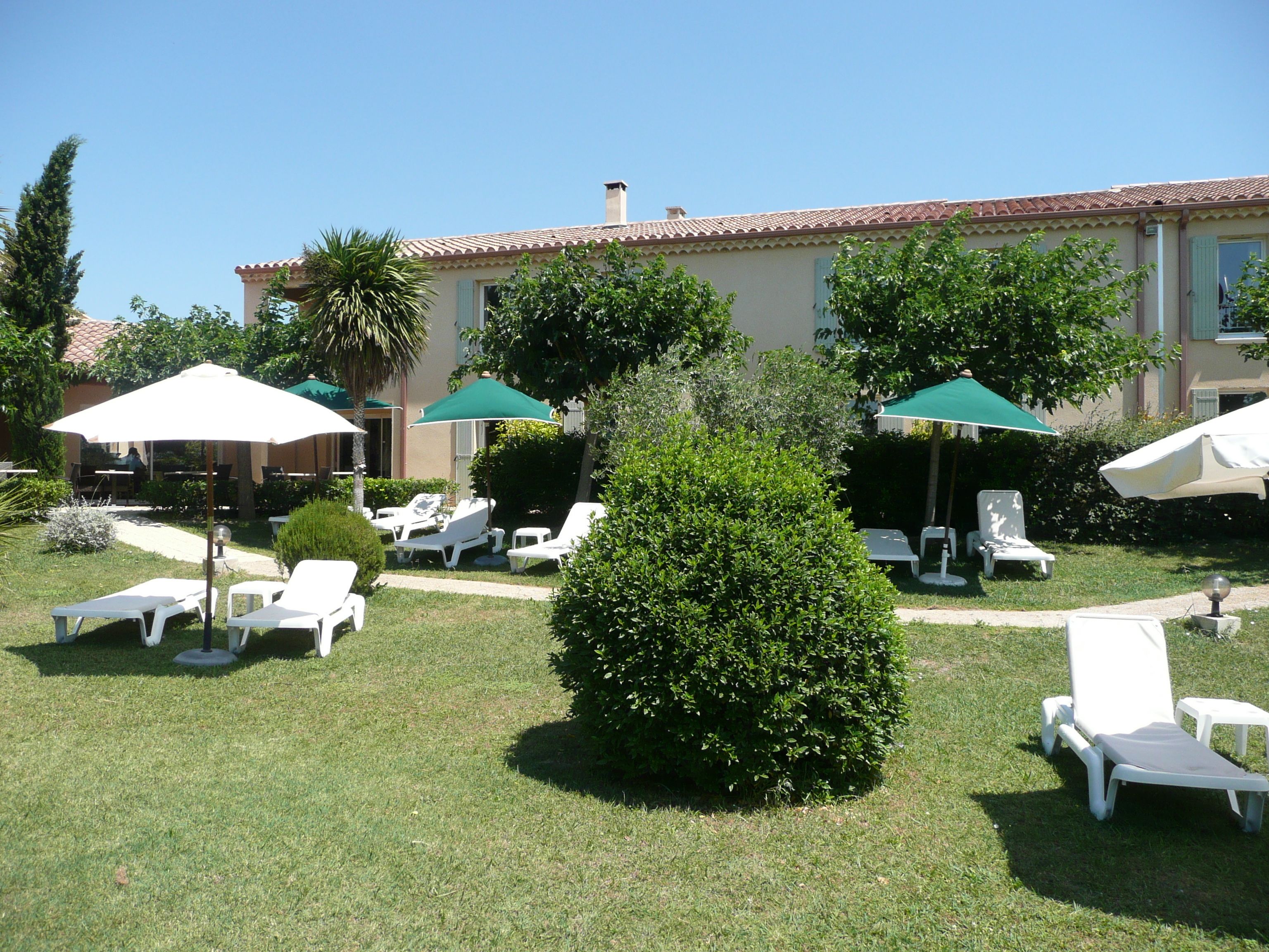 Un espace extérieur aménagé avec des fauteuils de jardin blancs et des parasols verts, entouré de pelouse et d’arbustes bien entretenus.