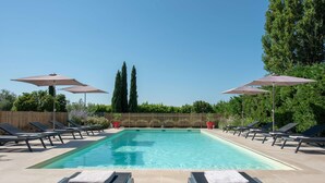 Une piscine rectangulaire à l’eau d’un bleu limpide, entourée de chaises longues et de parasols, avec une clôture en bois en arrière-plan.