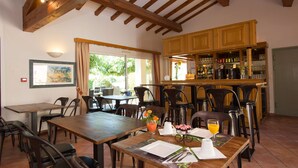 Un espace repas avec des tables et des chaises en bois, un bar avec des étagères remplies de bouteilles, et une vue sur la verdure à l’extérieur.