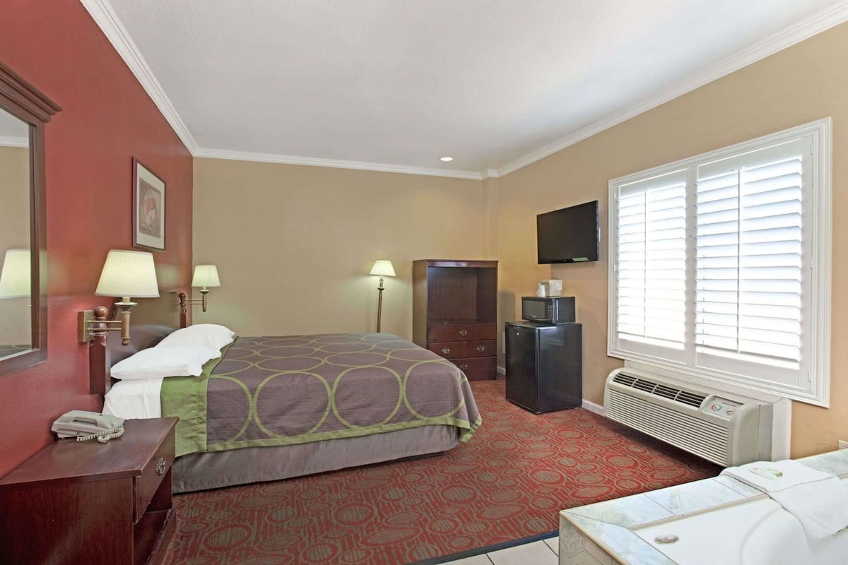 A hotel room with a bed, bedside lamps, a desk with a telephone, a television, a refrigerator, and a window with blinds.