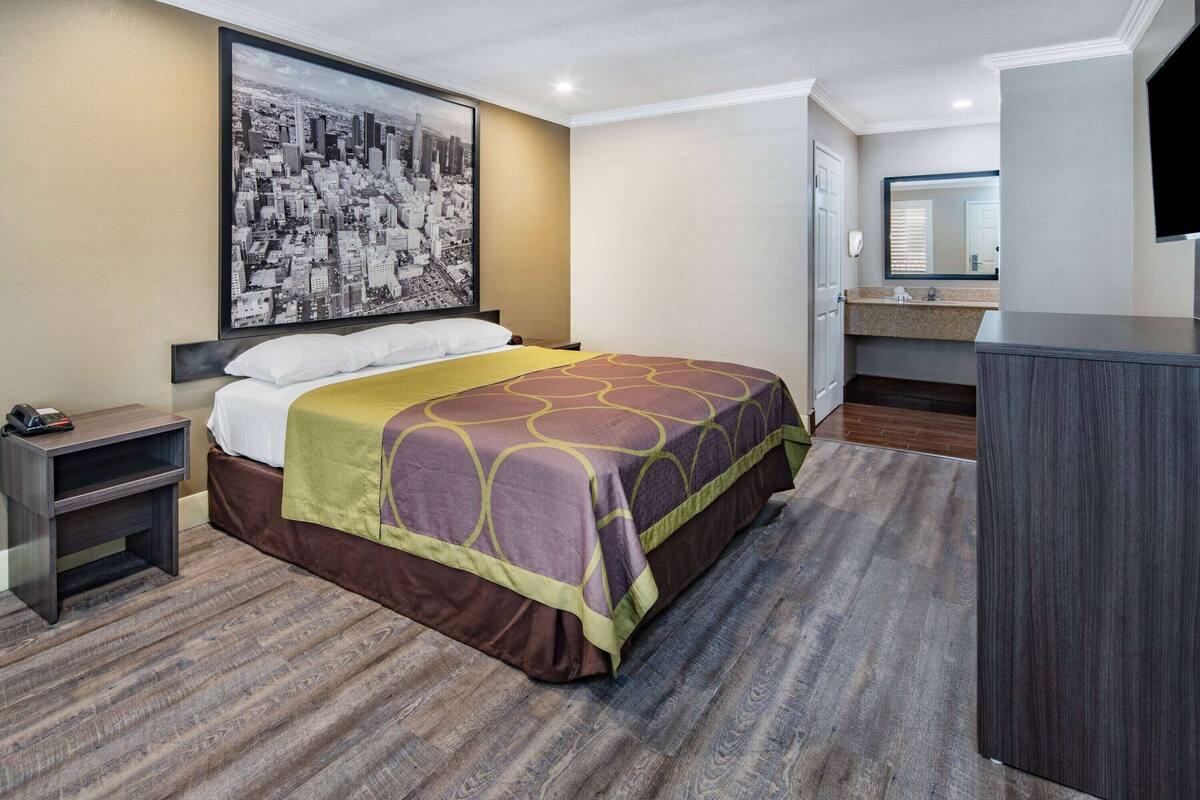 A hotel room with a bed, a nightstand, a large cityscape painting, a bathroom with a sink and mirror, and a flat-screen TV.