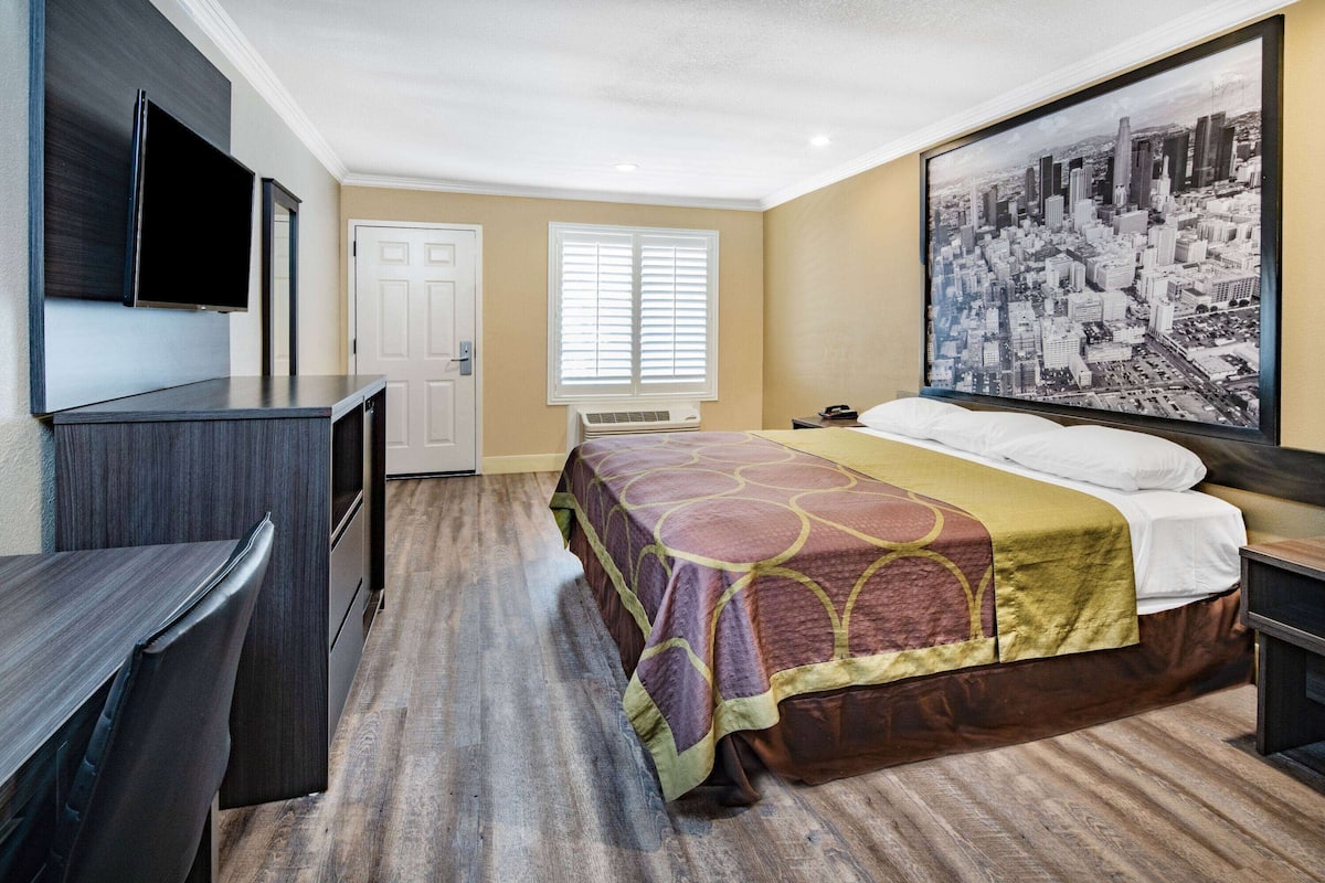 A hotel room with a bed, a desk, a television, and a large cityscape painting on the wall.