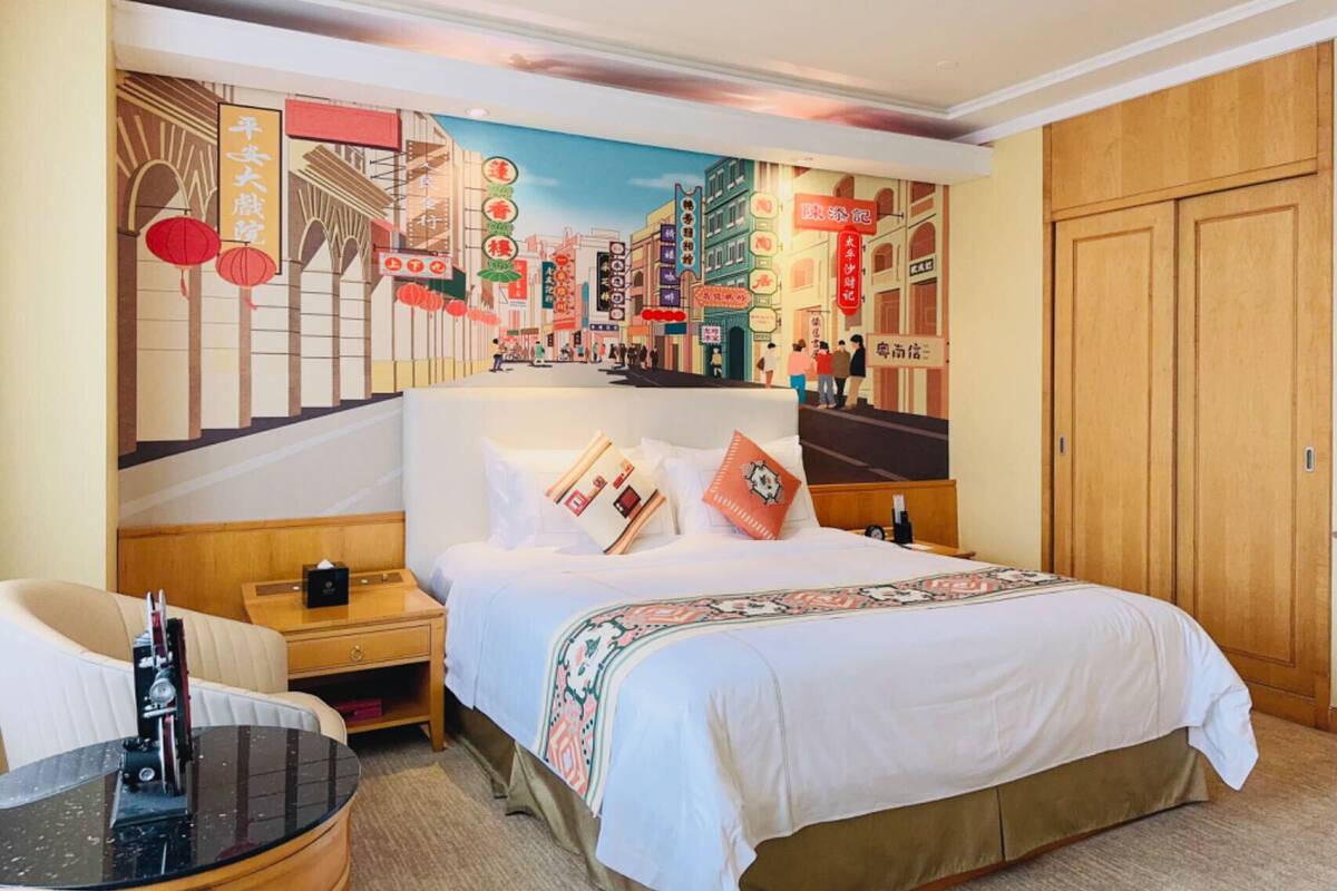 Une chambre d’hôtel avec un grand lit, une table de chevet, une chaise et une fresque murale représentant un paysage urbain.