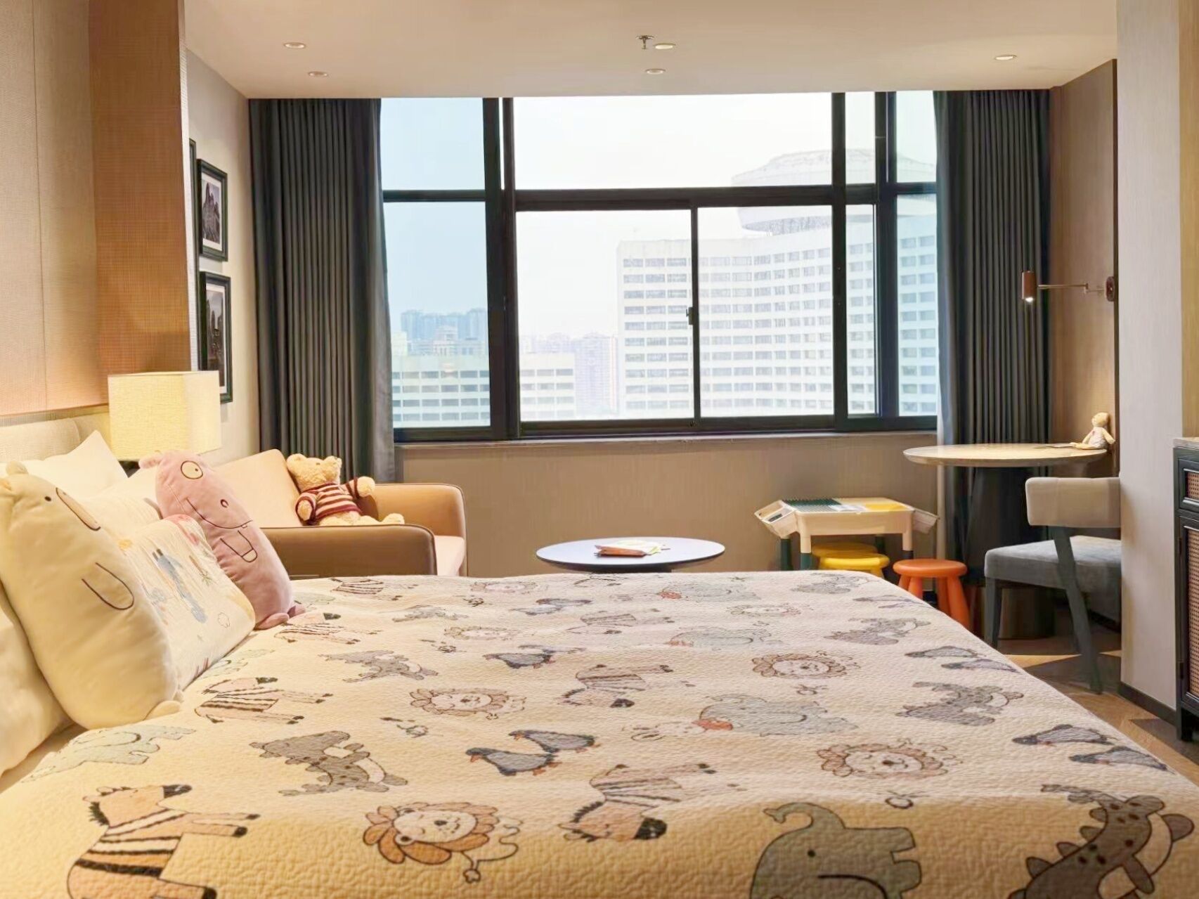 Habitación de hotel con una cama grande, un escritorio, una silla y vistas a la ciudad a través de amplias ventanas.