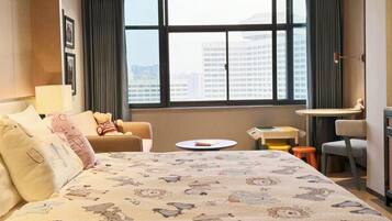 Habitación de hotel con una cama grande, un escritorio, una silla y vistas a la ciudad a través de amplias ventanas.