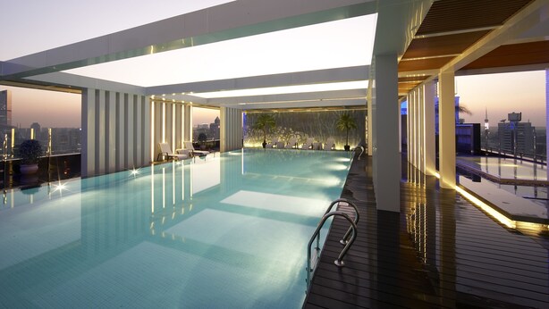 Piscina infinita en la azotea con vista a la ciudad, rodeada de arquitectura moderna y sillones de descanso.