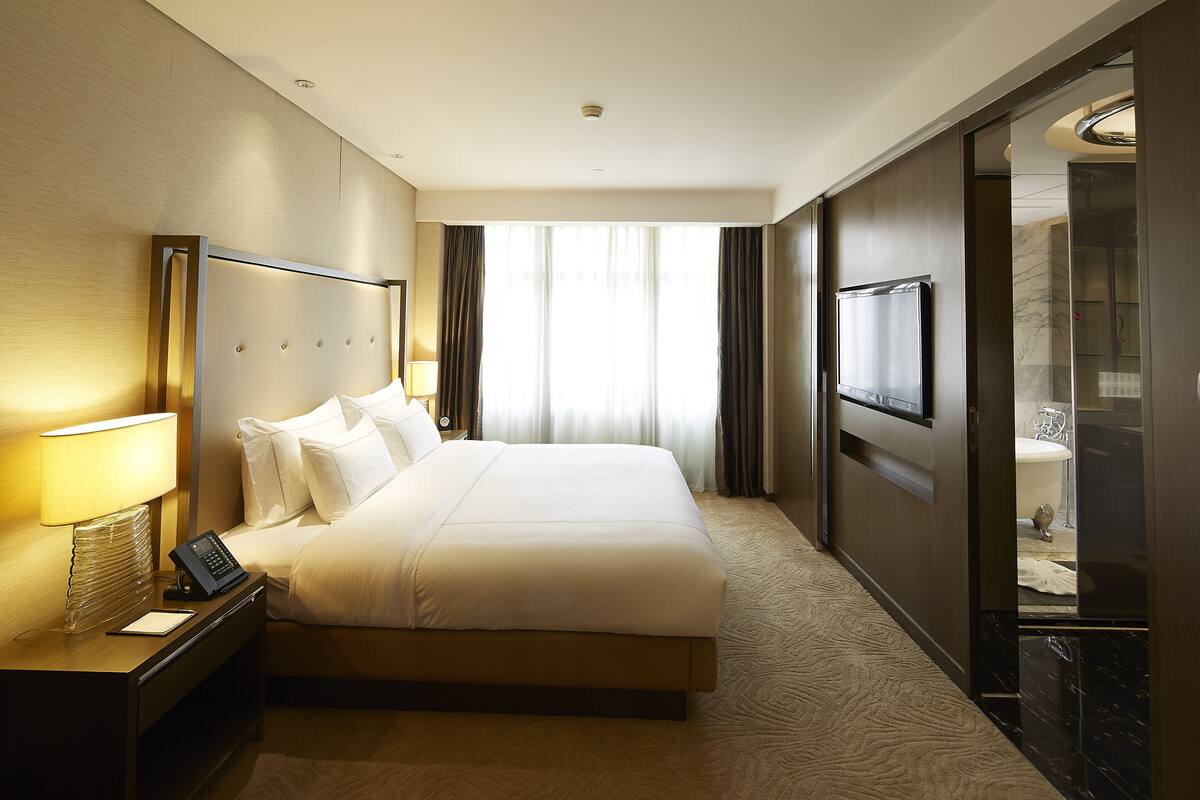 Une chambre d’hôtel avec un grand lit, une table de chevet avec une lampe, un téléviseur à écran plat et une vue sur la salle de bain à travers une porte en verre.