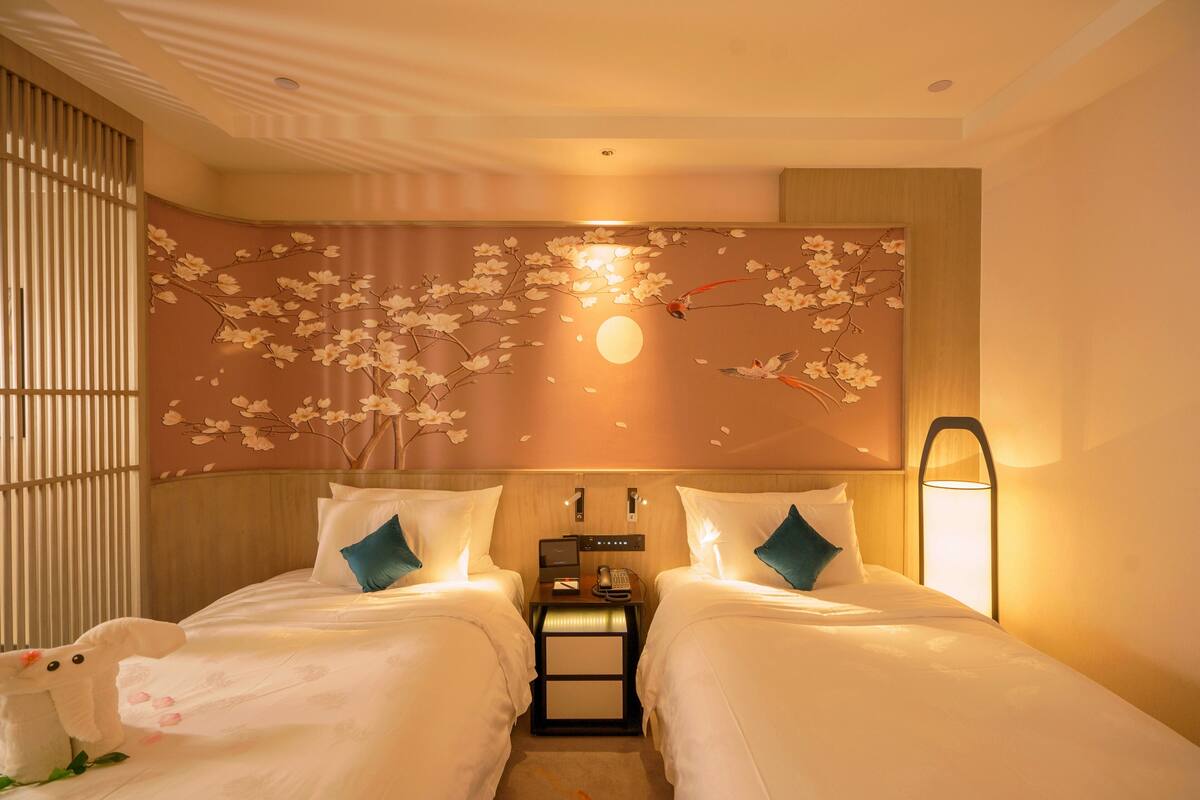 Une chambre d’hôtel avec deux lits, une table de chevet, une lampe fixée au mur et un mur décoratif orné de motifs de fleurs de cerisier et d’une lune.