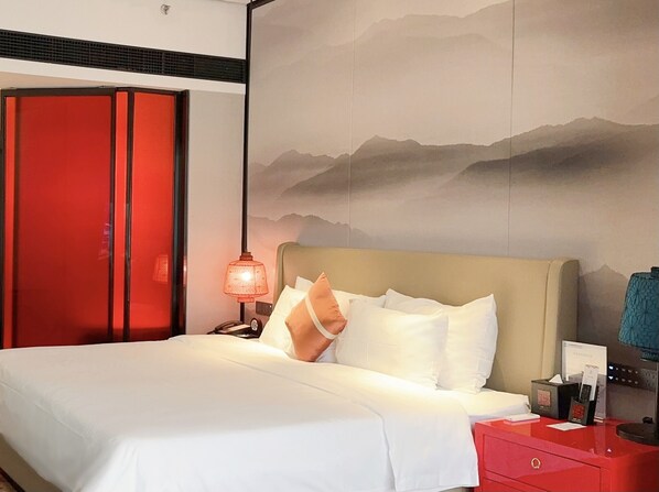 Habitación de hotel con cama, mesita de noche, lámpara y un cuadro de un paisaje montañoso.
