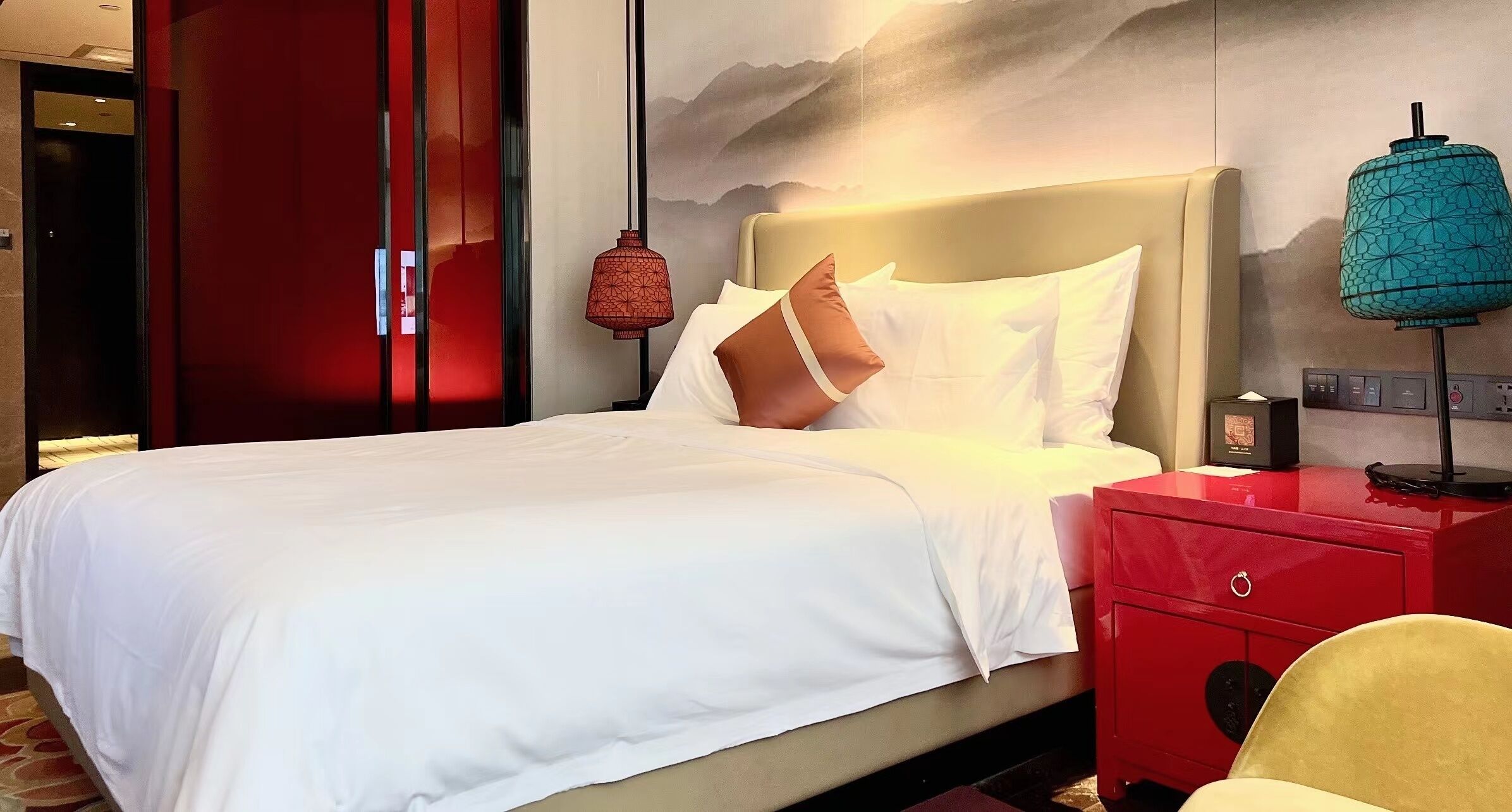 A hotel room with a bed, a red nightstand, a lamp, and a wall with a mountain painting.