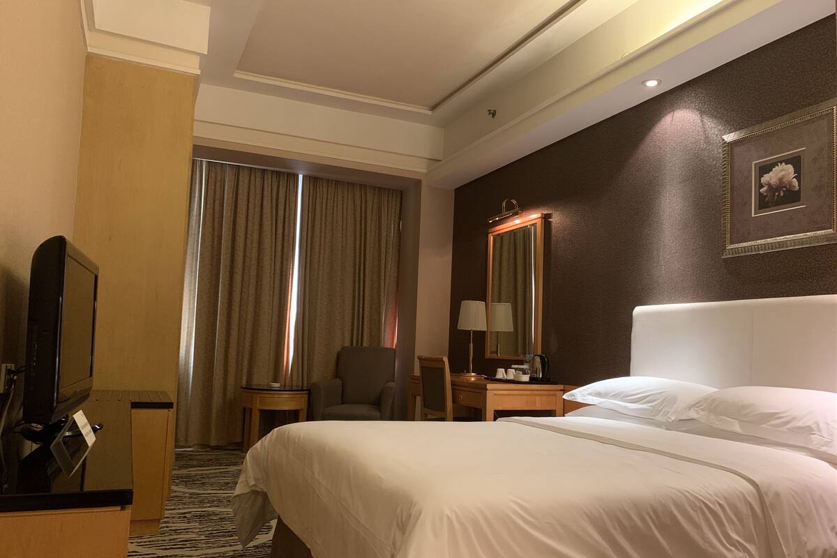 Une chambre d’hôtel avec un grand lit, un bureau avec un ordinateur, une chaise, un miroir et une fenêtre avec des rideaux.