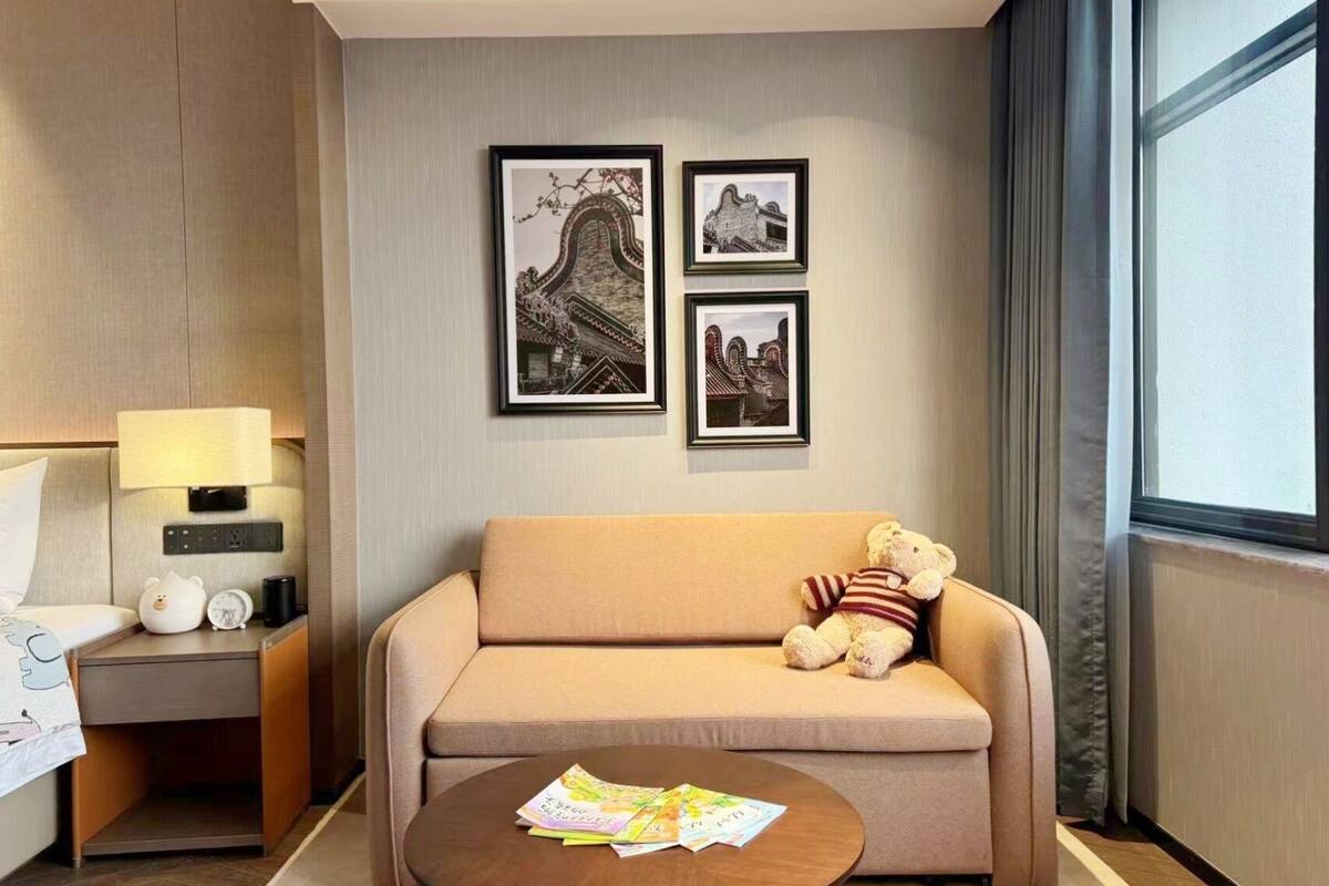 Une chambre d’hôtel avec un canapé, une petite table avec des magazines, une lampe, une table de chevet avec une horloge et un ours en peluche, et trois tableaux encadrés accrochés au mur.