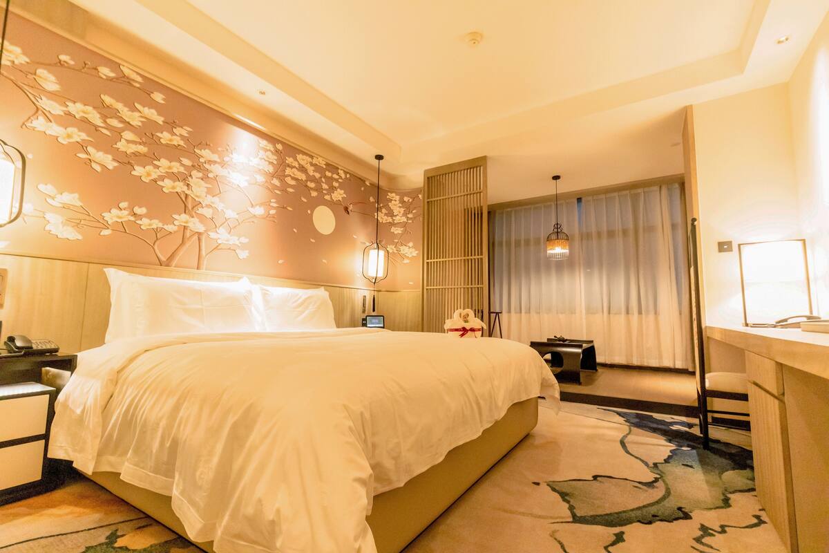 Une chambre d’hôtel avec un grand lit, un bureau, une chaise, une table de chevet, une lampe et un mur décoratif orné de motifs floraux.