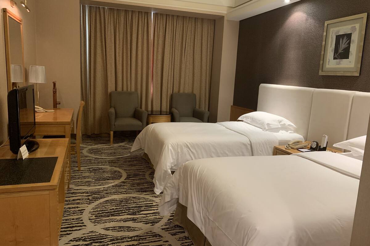 Une chambre d’hôtel avec deux lits, un bureau équipé d’un ordinateur et d’un téléphone, une chaise, une petite table et une grande fenêtre avec des rideaux.