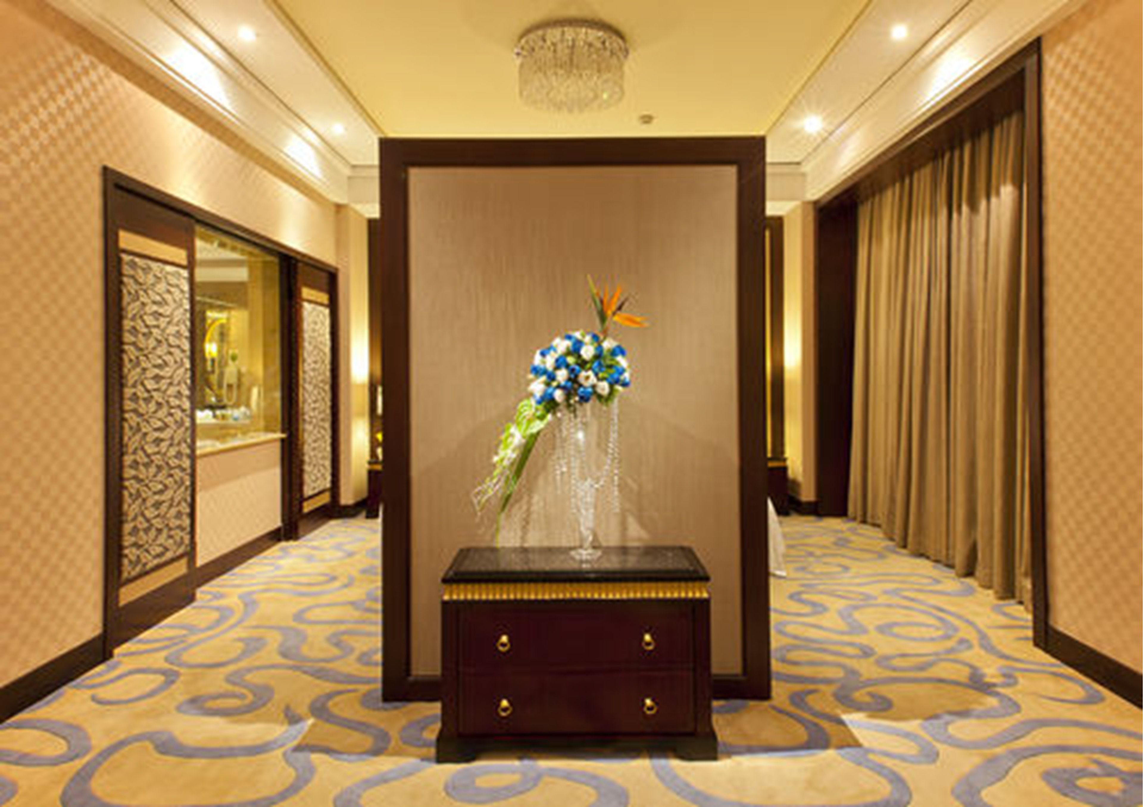 A hotel lobby with a decorative vase of flowers on a wooden table, patterned carpet, and a chandelier.