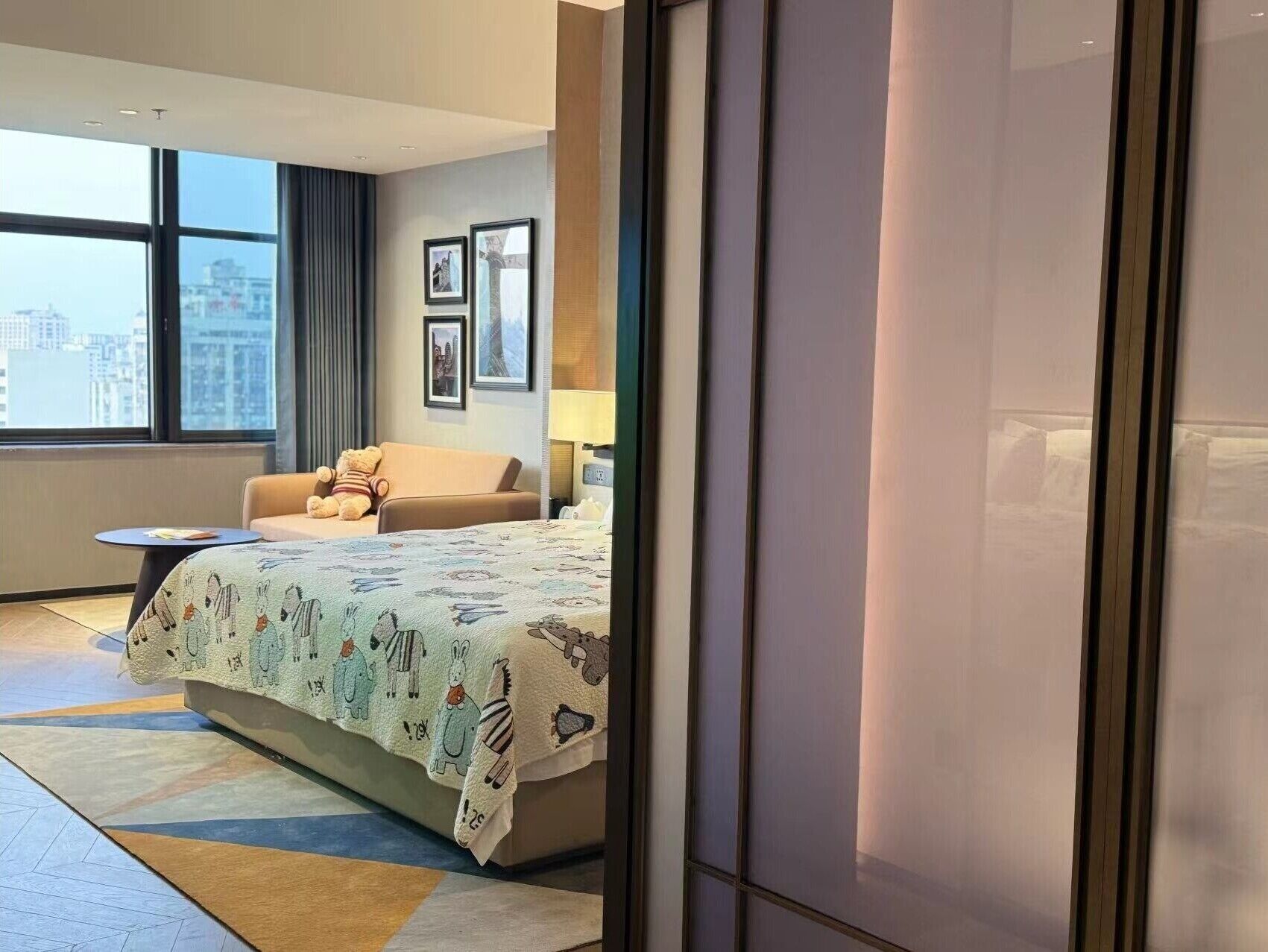 A modern hotel room with a large bed, a sofa, a small table, and a large window offering a city view.