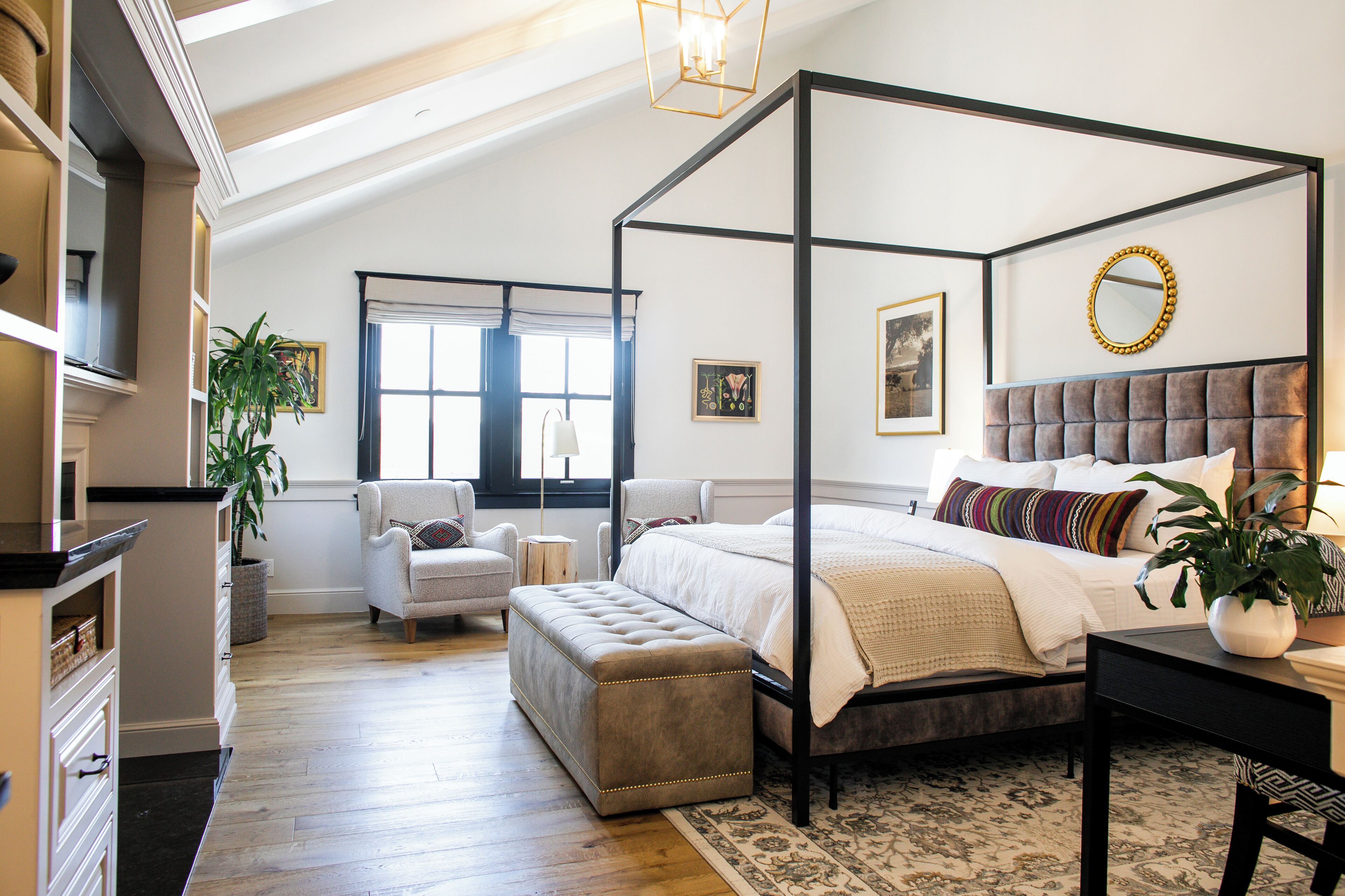 A bedroom with a four-poster bed, a tufted headboard, a bedside table, a chair, a bench, a potted plant, and a window with curtains.