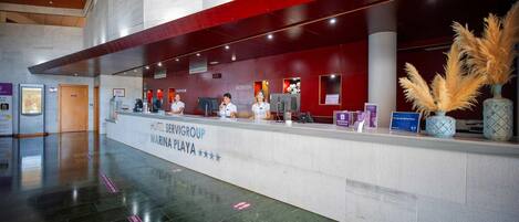 A hotel reception area with a modern design, featuring a reception desk, staff, and a decorative plant.