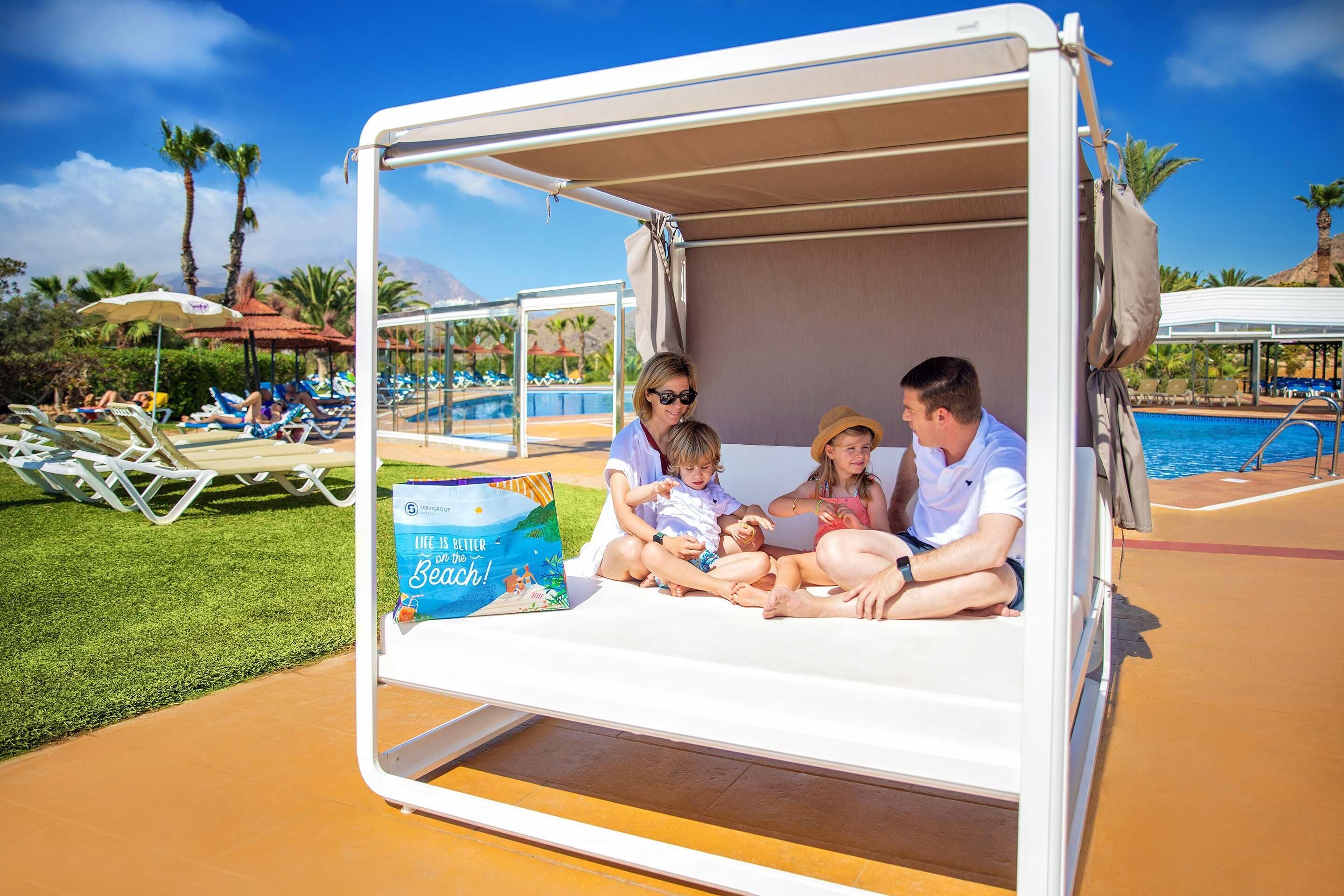 A family of four seated in a sun lounger with a pool view, a beach-themed book, and a poolside setting with lounge chairs and umbrellas.