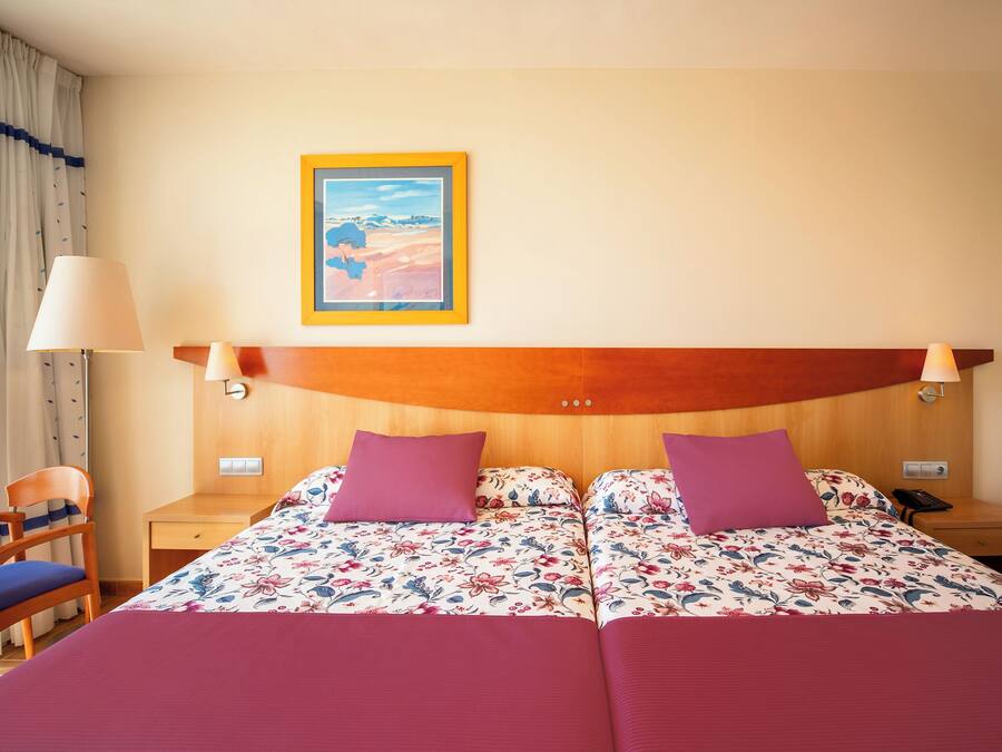Cama doble con ropa de cama floral, cabecera de madera, dos mesitas de noche, una silla y una mesita con lámpara.