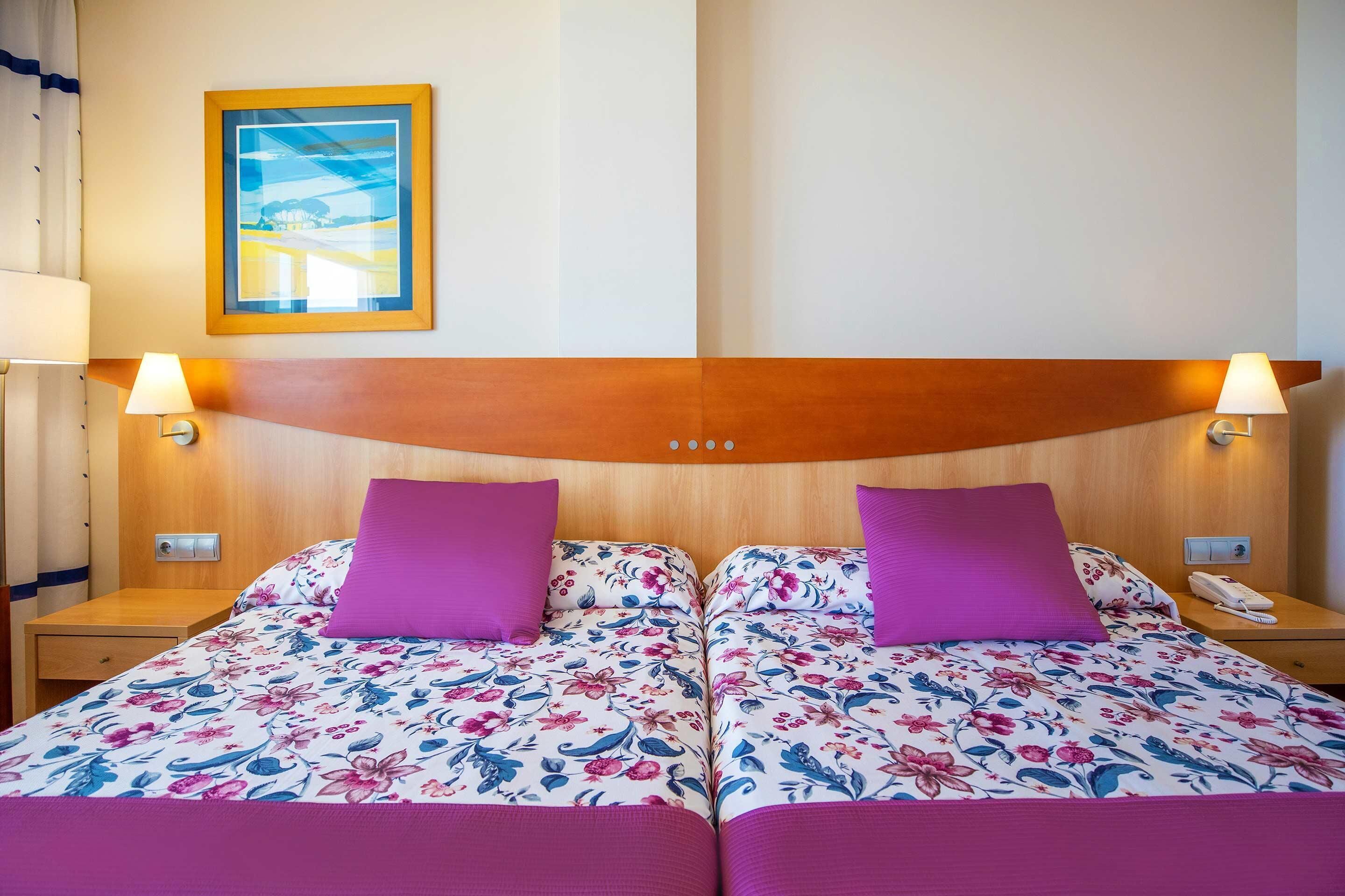Un lit double avec une literie à motifs floraux et deux oreillers violets, une tête de lit en bois, des lampes de chevet, une table de chevet et un tableau encadré au mur.