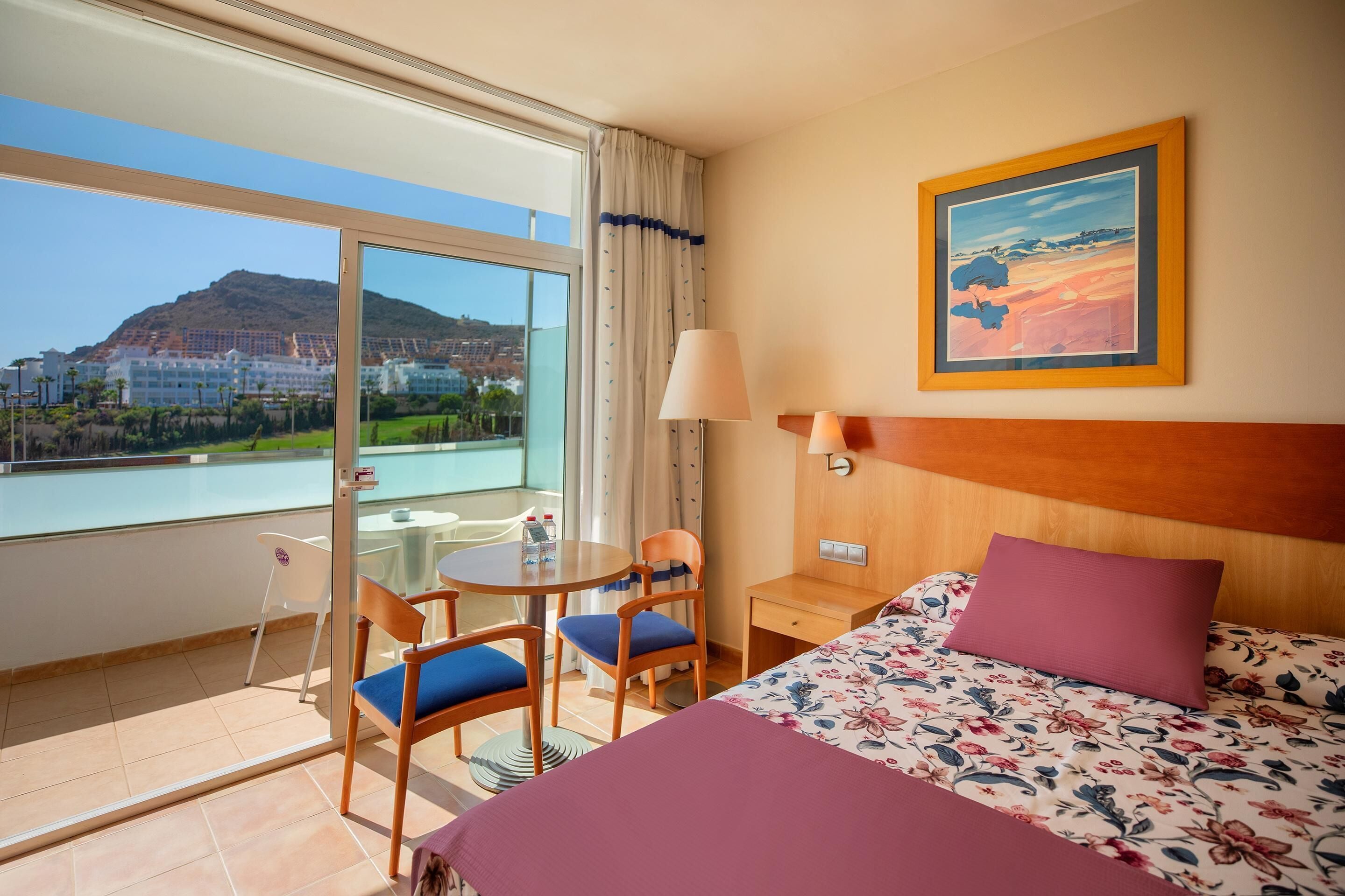 A hotel room with a bed, a table, two chairs, a lamp, and a painting.