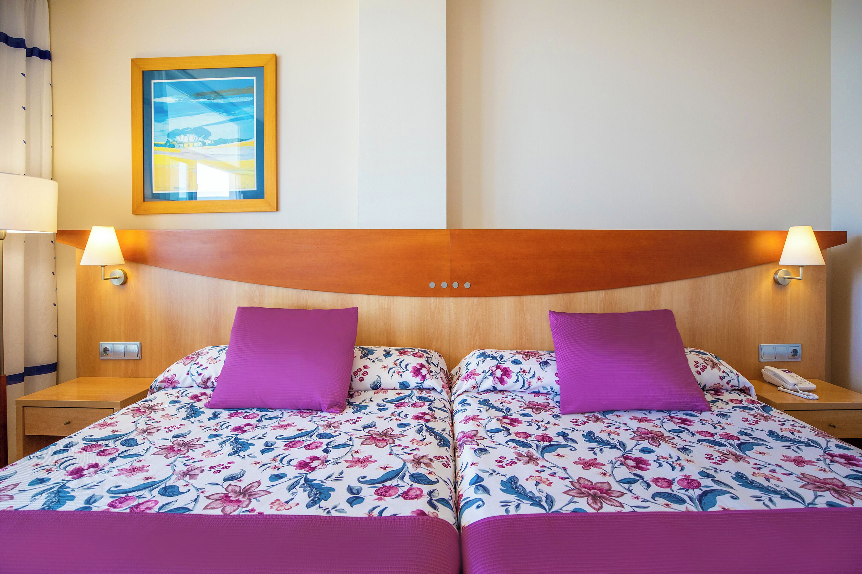 A double bed with floral bedding and two purple pillows, a wooden headboard, bedside lamps, a nightstand, a framed picture on the wall, and a phone on the nightstand.