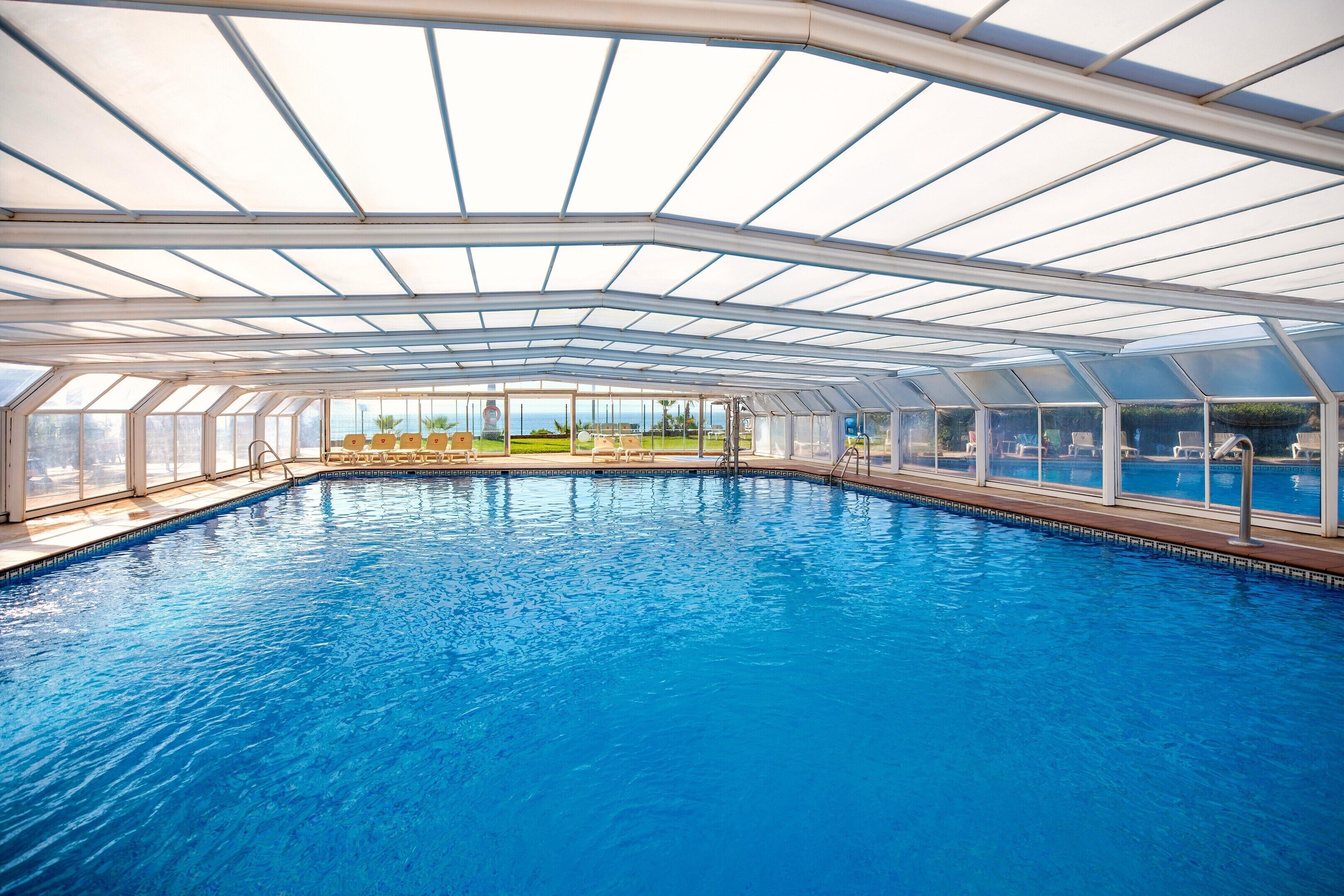 A large indoor swimming pool with a glass ceiling, surrounded by lounge chairs and a view of the outdoors.