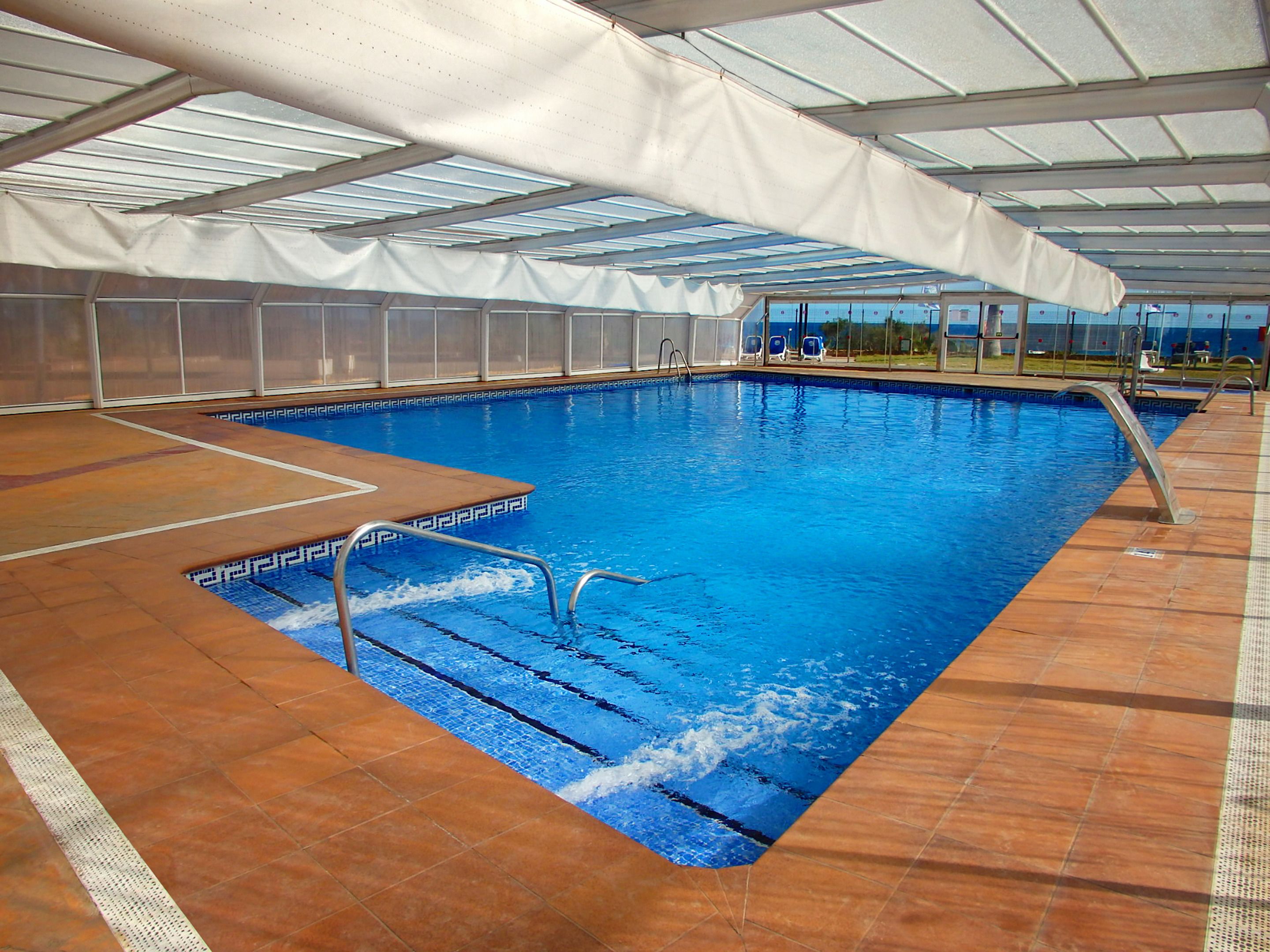 Una piscina interior con agua azul cristalina, una terraza de madera y una zona cubierta con techo.