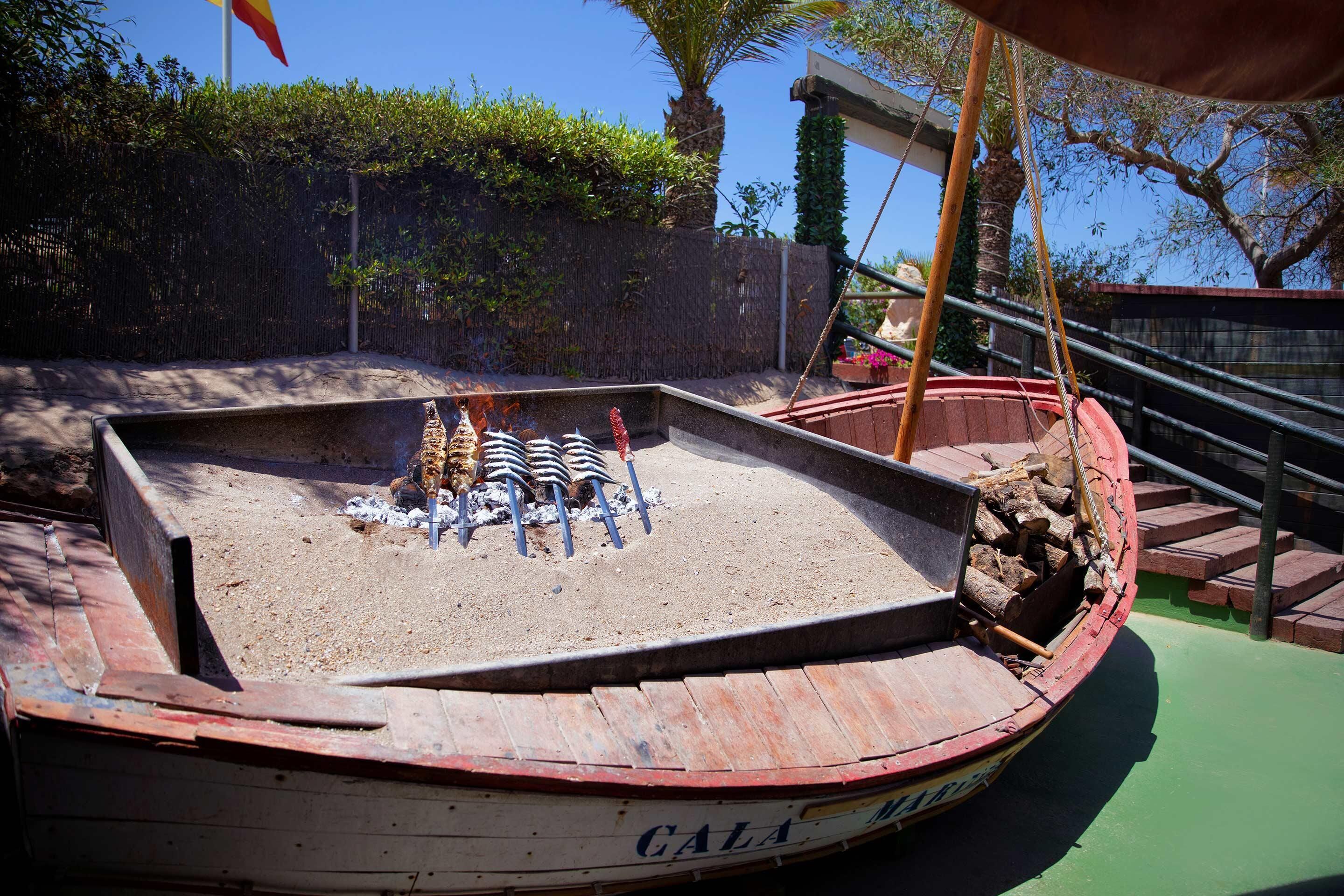 A boat with the name "CALA" on its side, containing logs and a grill with skewers of food.
