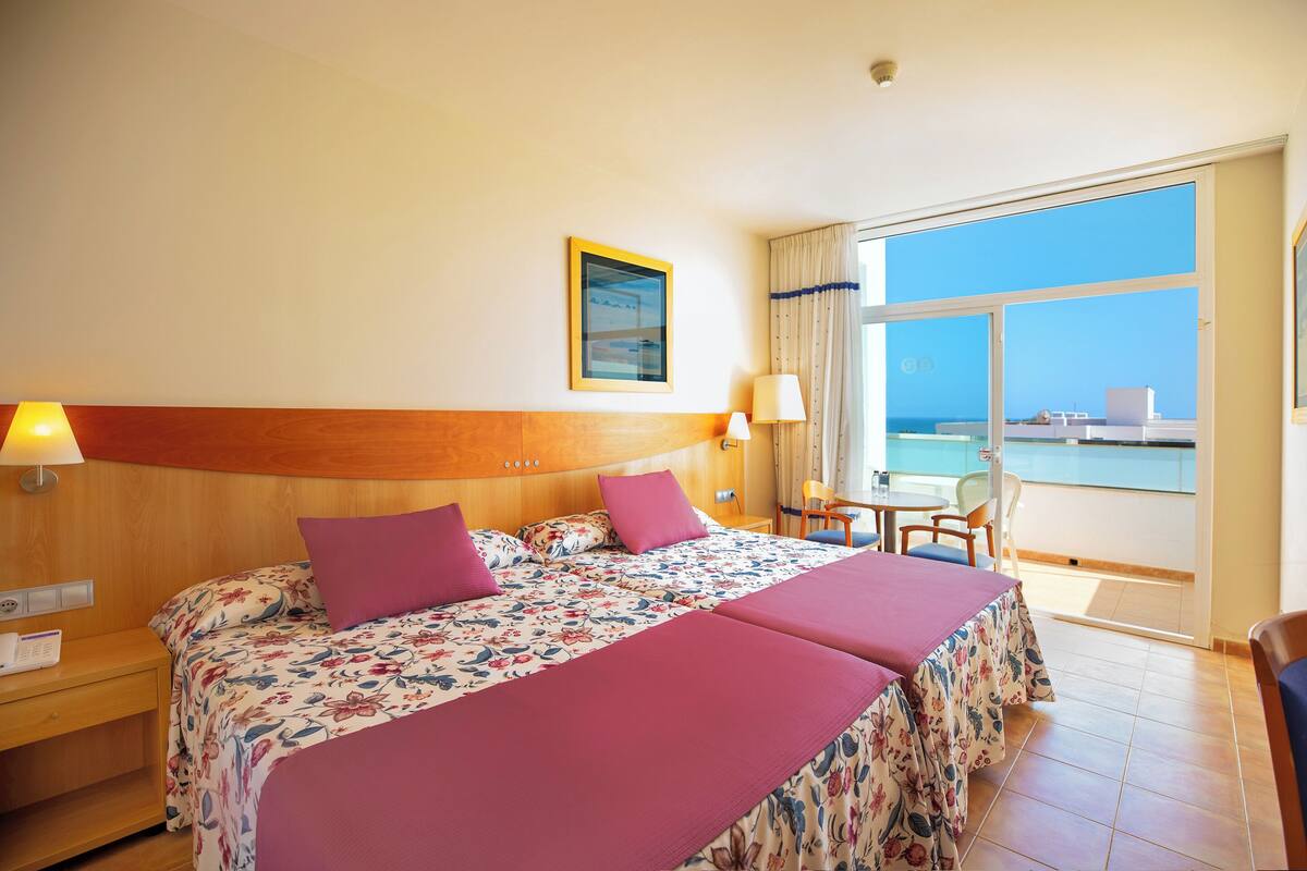 Quarto de hotel com duas camas, varanda com vista para o mar e uma pequena mesa com cadeiras.