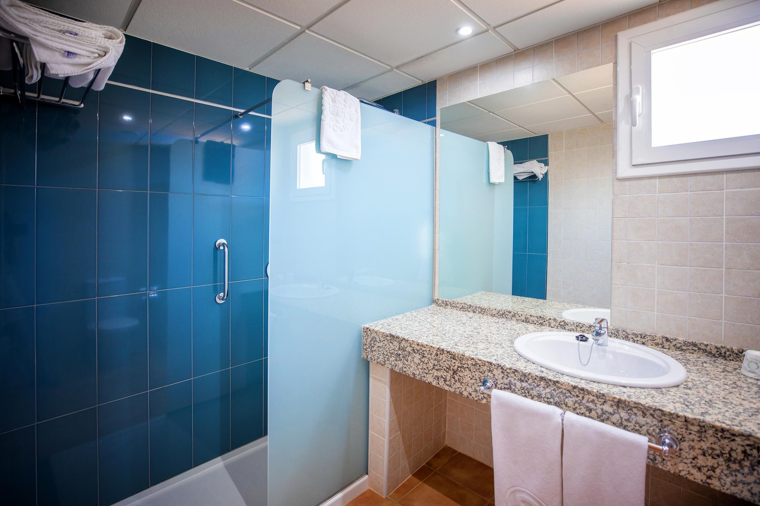 Un baño con pared de ducha de azulejos azules, lavamanos de encimera de granito y un espejo encima.
