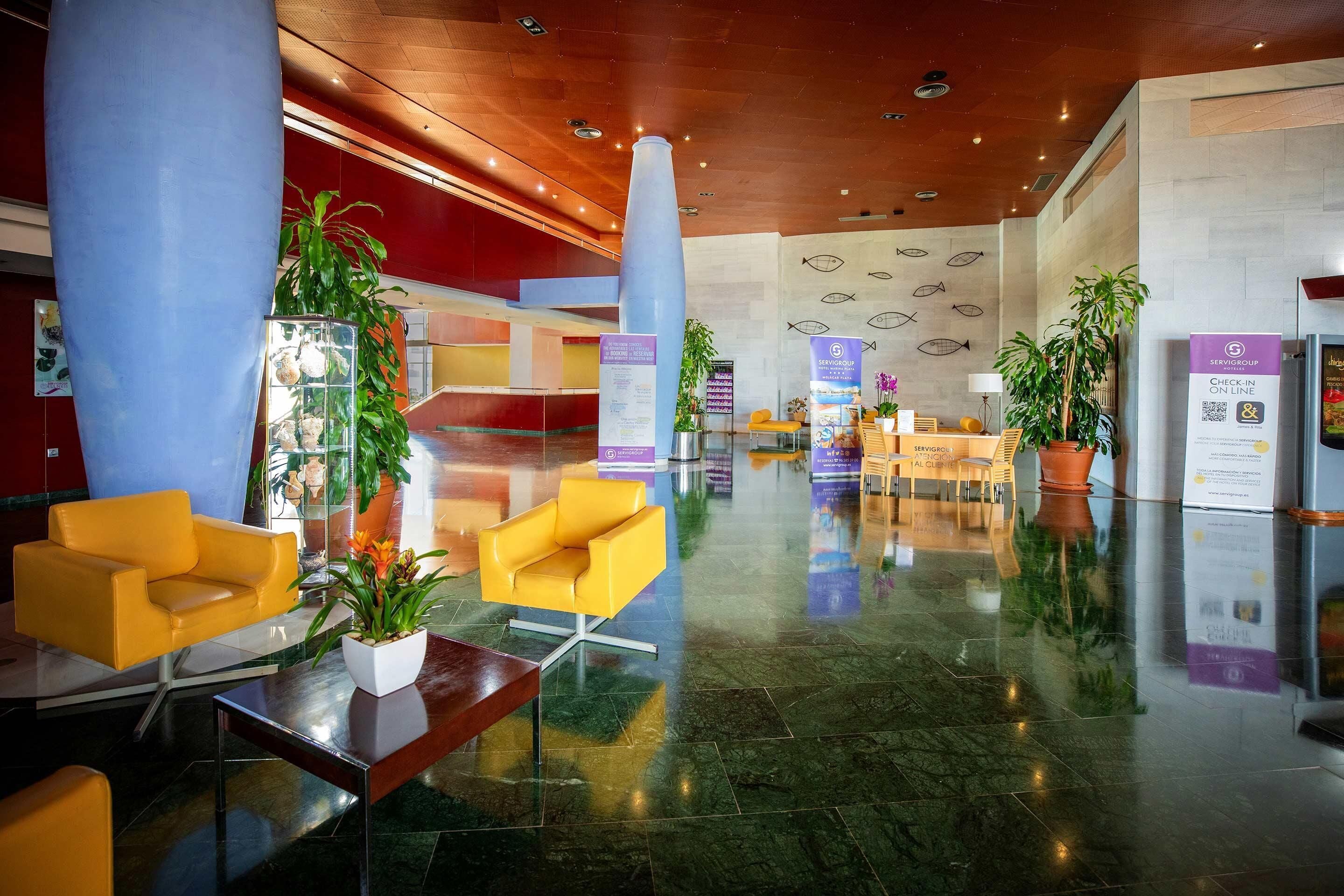 A spacious lobby with a modern design, featuring a reception desk, seating area with yellow chairs, and potted plants.