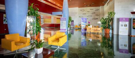 A spacious lobby with a modern design, featuring a reception desk, seating area with yellow chairs, and potted plants.